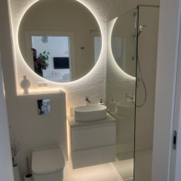 Artur Wolak Bud - Nowoczesna, jasna łazienka z okrągłym lustrem podświetlanym LED, białą armaturą, kabiną prysznicową ze szkła i płytkami imitującymi kamień na ścianach.