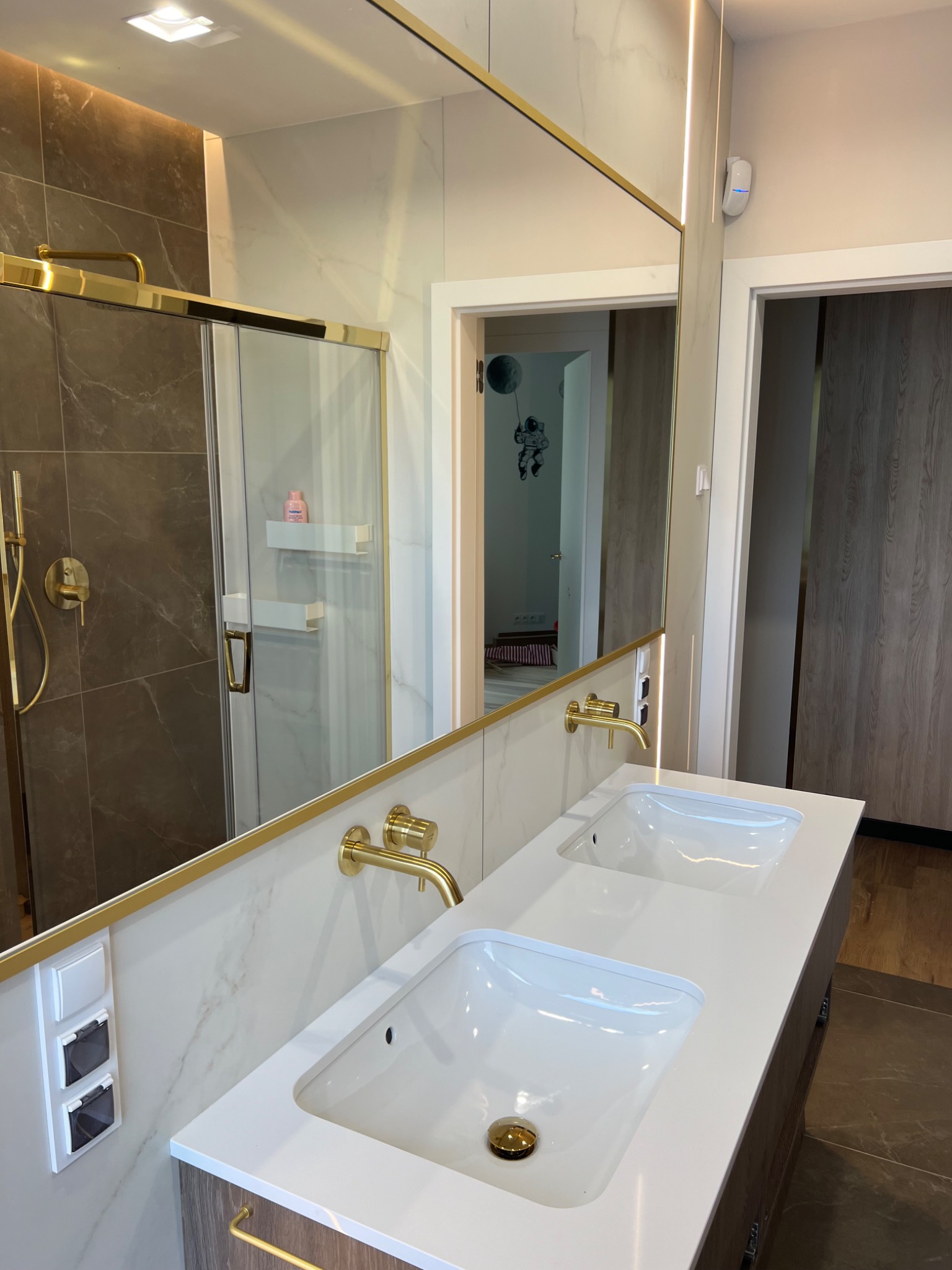 Nowoczesna łazienka z podwójną umywalką, złotymi bateriami i kabiną prysznicową ze złotymi detalami, odbicie wnętrza w lustrze.