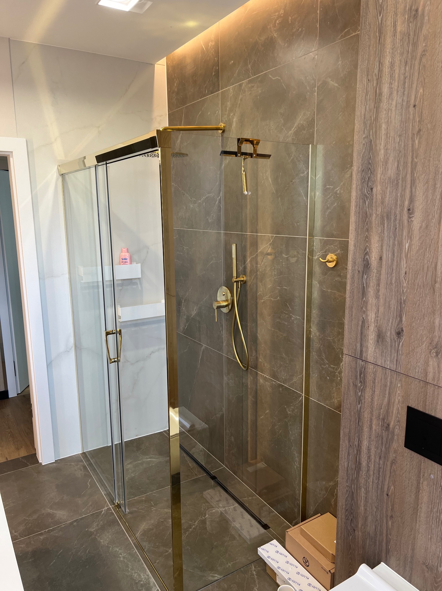 Nowoczesna kabina prysznicowa z przesuwnymi drzwiami, złotymi okuciami i szarymi płytkami imitującymi marmur, widoczne pudełka po armaturze na podłodze.