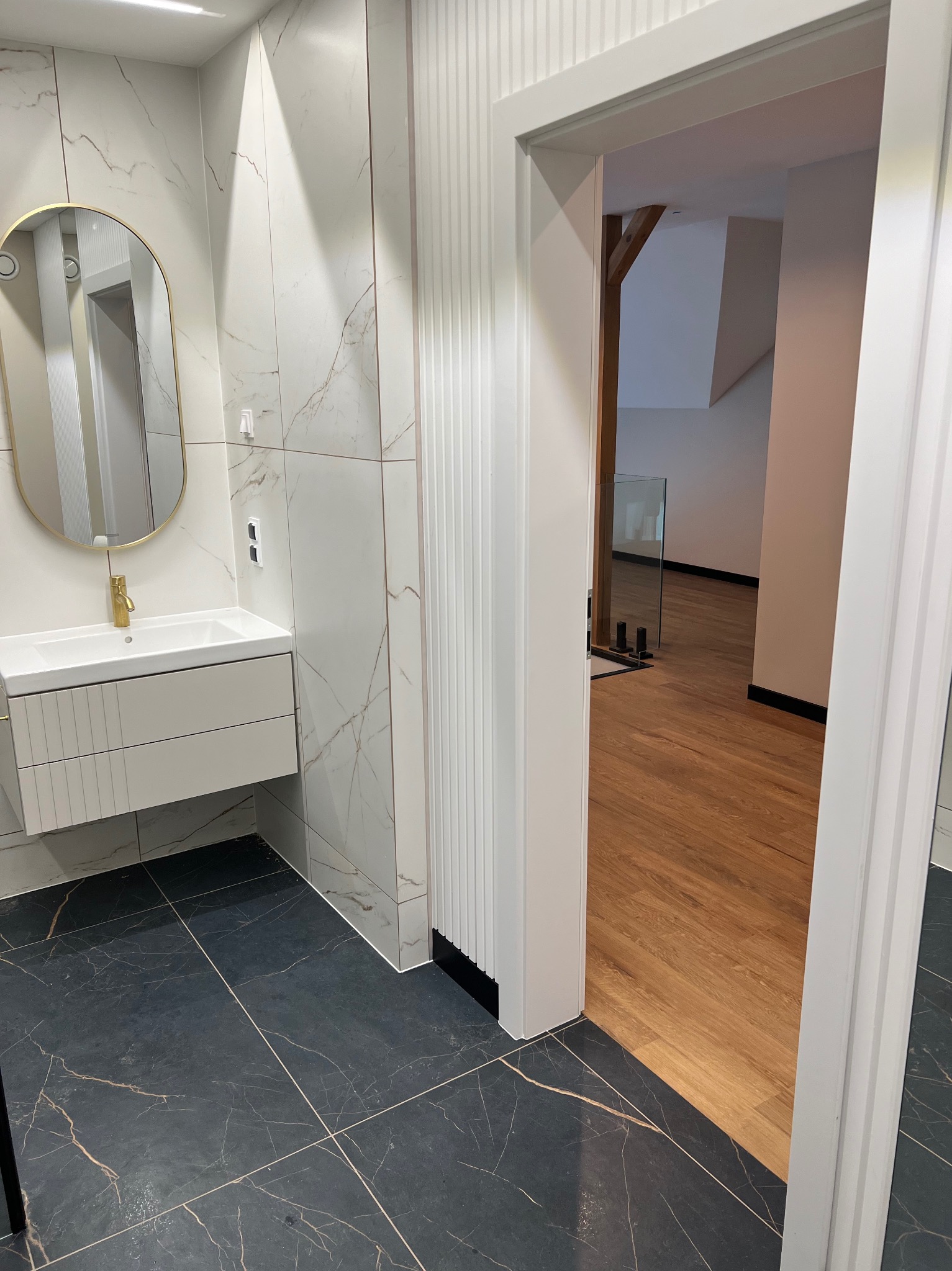 Nowoczesna łazienka z umywalką nablatową, złotą armaturą i lustrem w złotej ramie, czarna podłoga z marmurkowym wzorem, widok na drewnianą podłogę i elementy konstrukcyjne przez otwarte drzwi.
