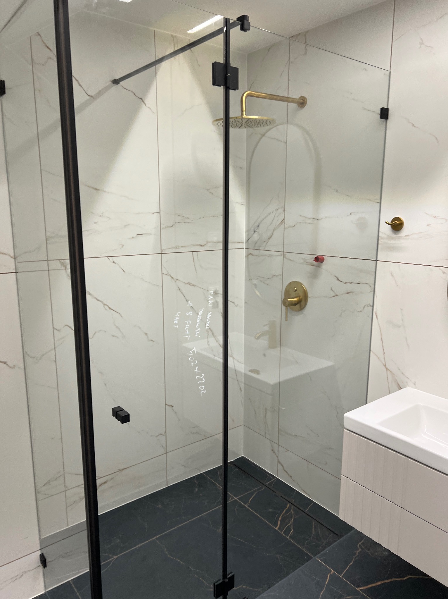 Nowoczesna kabina prysznicowa z czarnymi ramami i złotą armaturą na tle białych płytek imitujących marmur i ciemnej podłogi. Na szkle widoczne odręczne notatki.