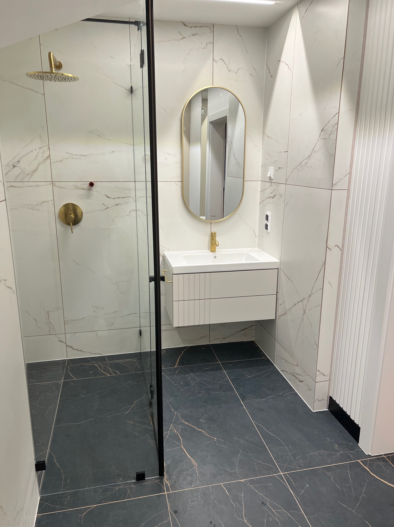 Nowoczesna łazienka z kabiną prysznicową typu walk-in z czarnymi profilami, umywalką nablatową z owalnym lustrem w złotej ramie i płytkami imitującymi marmur na ścianach i podłodze.