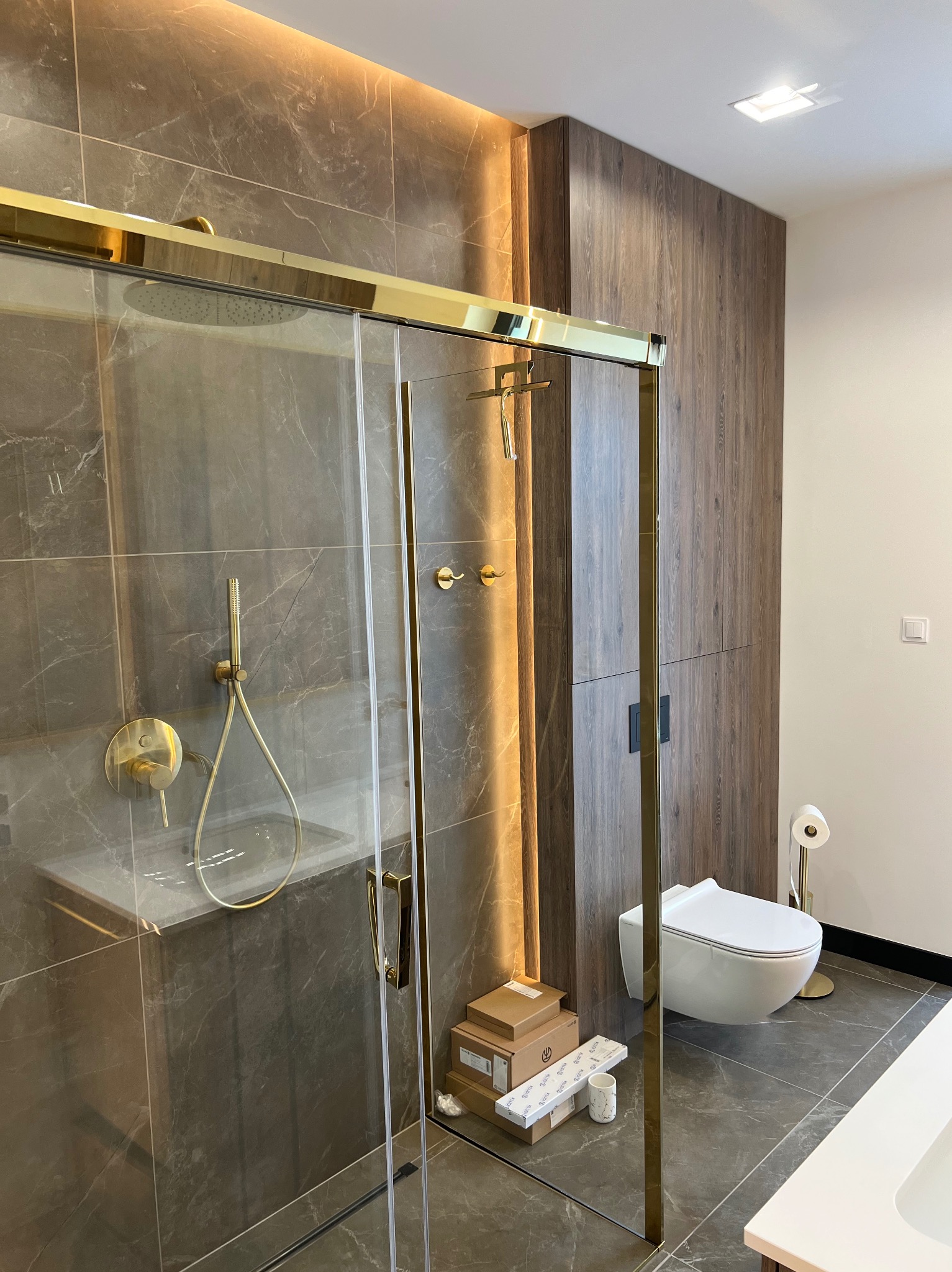 Nowoczesna łazienka z kabiną prysznicową ze złotymi detalami, marmurowe płytki, podwieszana toaleta i drewniana zabudowa.