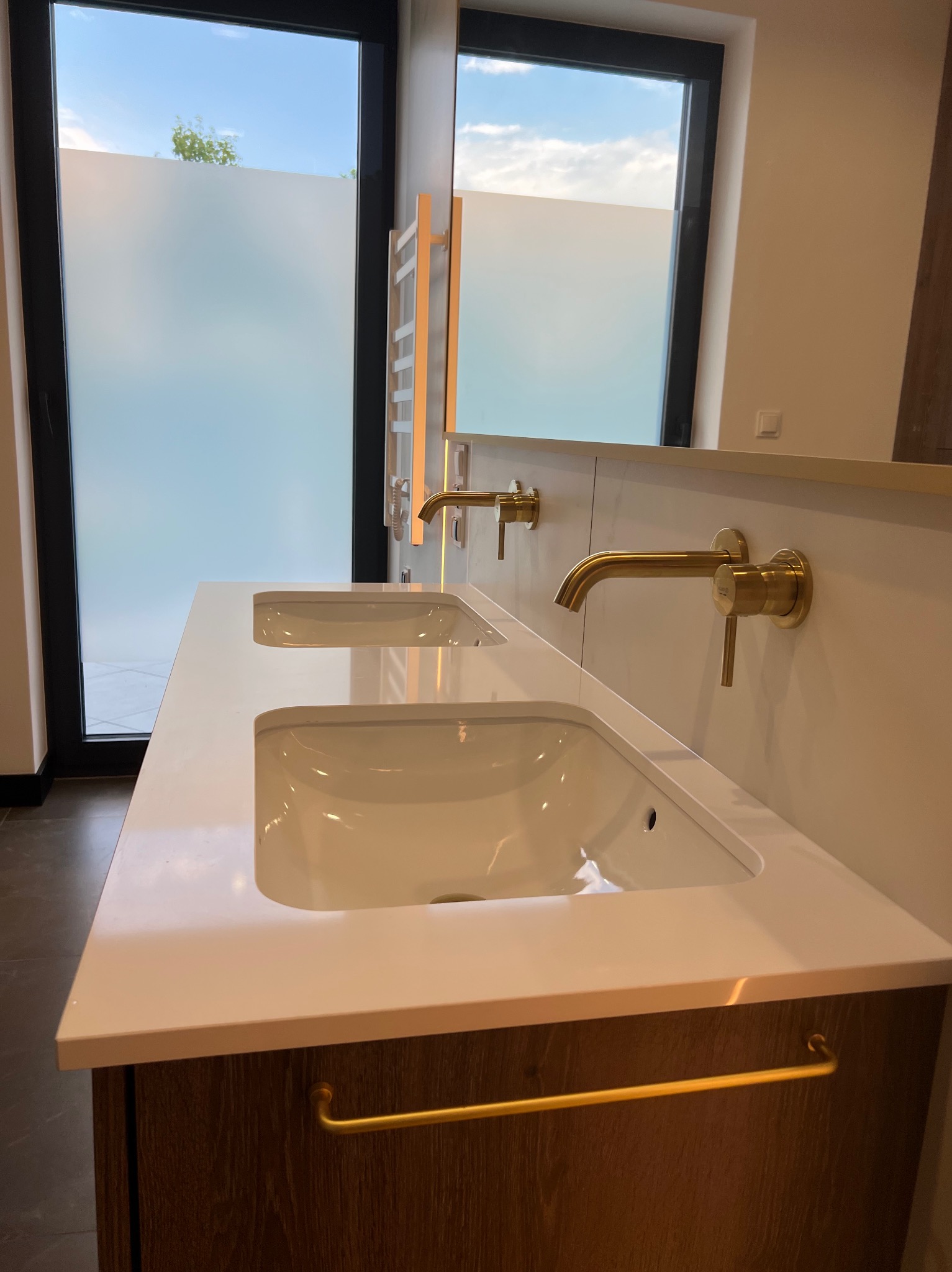 Nowoczesna łazienka z podwójną umywalką nablatową, złotą armaturą i szafką z drewnopodobnym wykończeniem, okno z matową folią w tle.