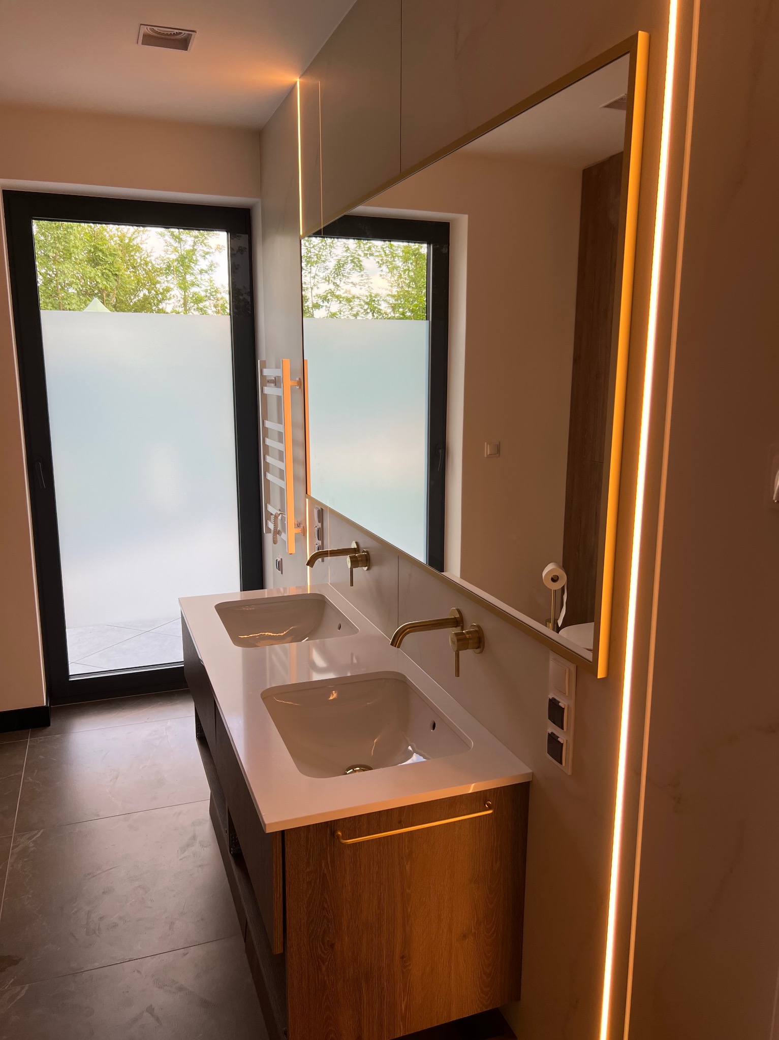 Nowoczesna łazienka z podwójną umywalką, złotymi bateriami i podświetlanym lustrem. Widoczne duże okno z matową szybą i grzejnik drabinkowy.