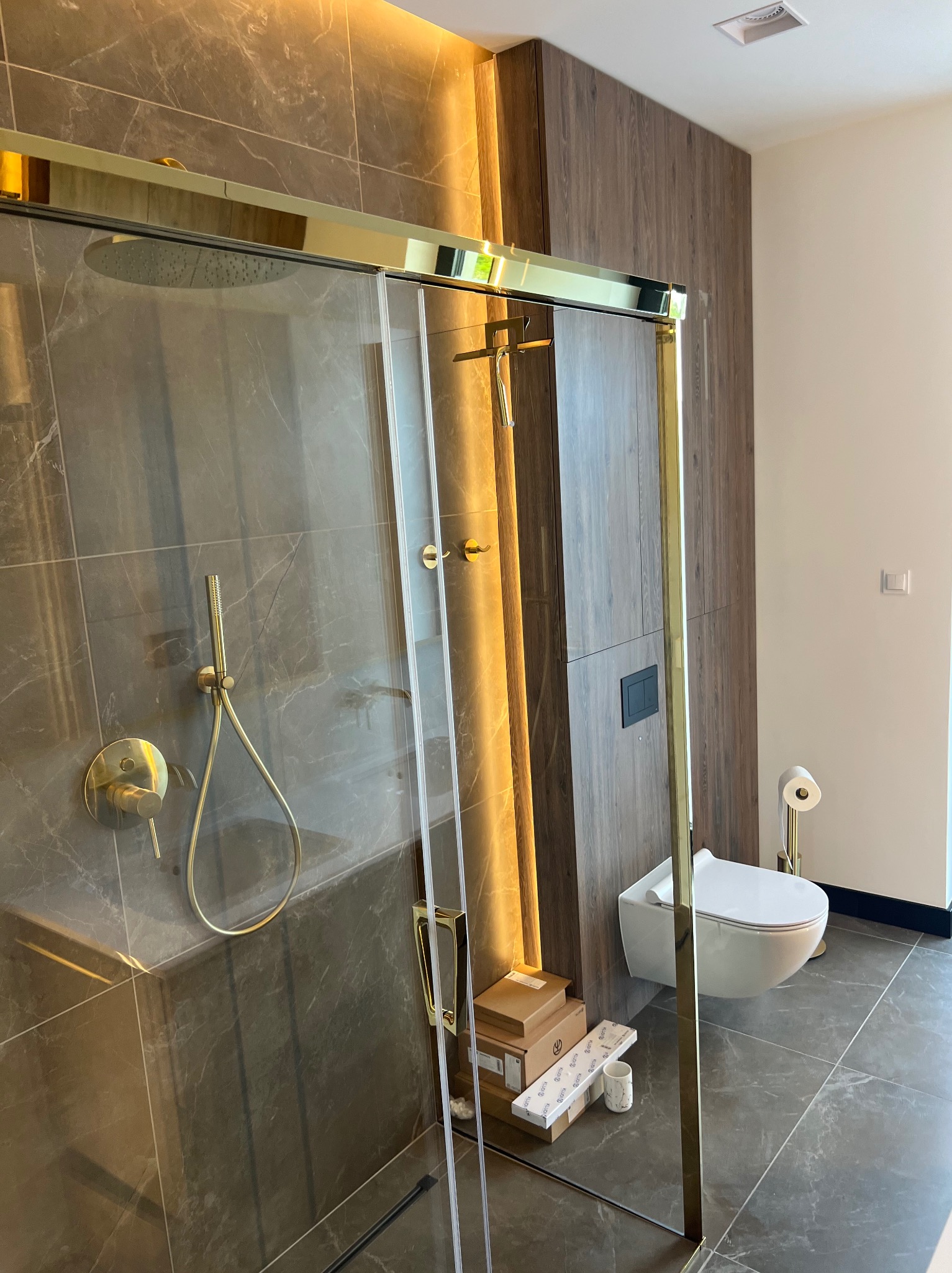 Nowoczesna łazienka z kabiną prysznicową w złotej ramie, marmurowymi płytkami, wiszącą toaletą i elementami złotego osprzętu, z pudełkami po akcesoriach na podłodze.