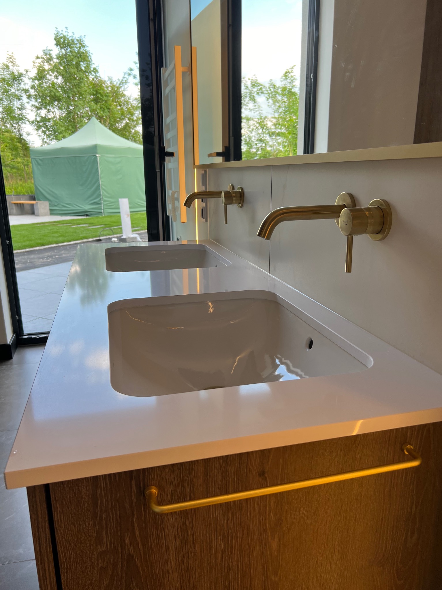 Nowoczesna łazienka z podwójną umywalką wpuszczoną w biały blat, drewniana szafka pod umywalką z złotym relingiem na ręczniki, złote baterie, widok na zielony trawnik i namiot przez okno.