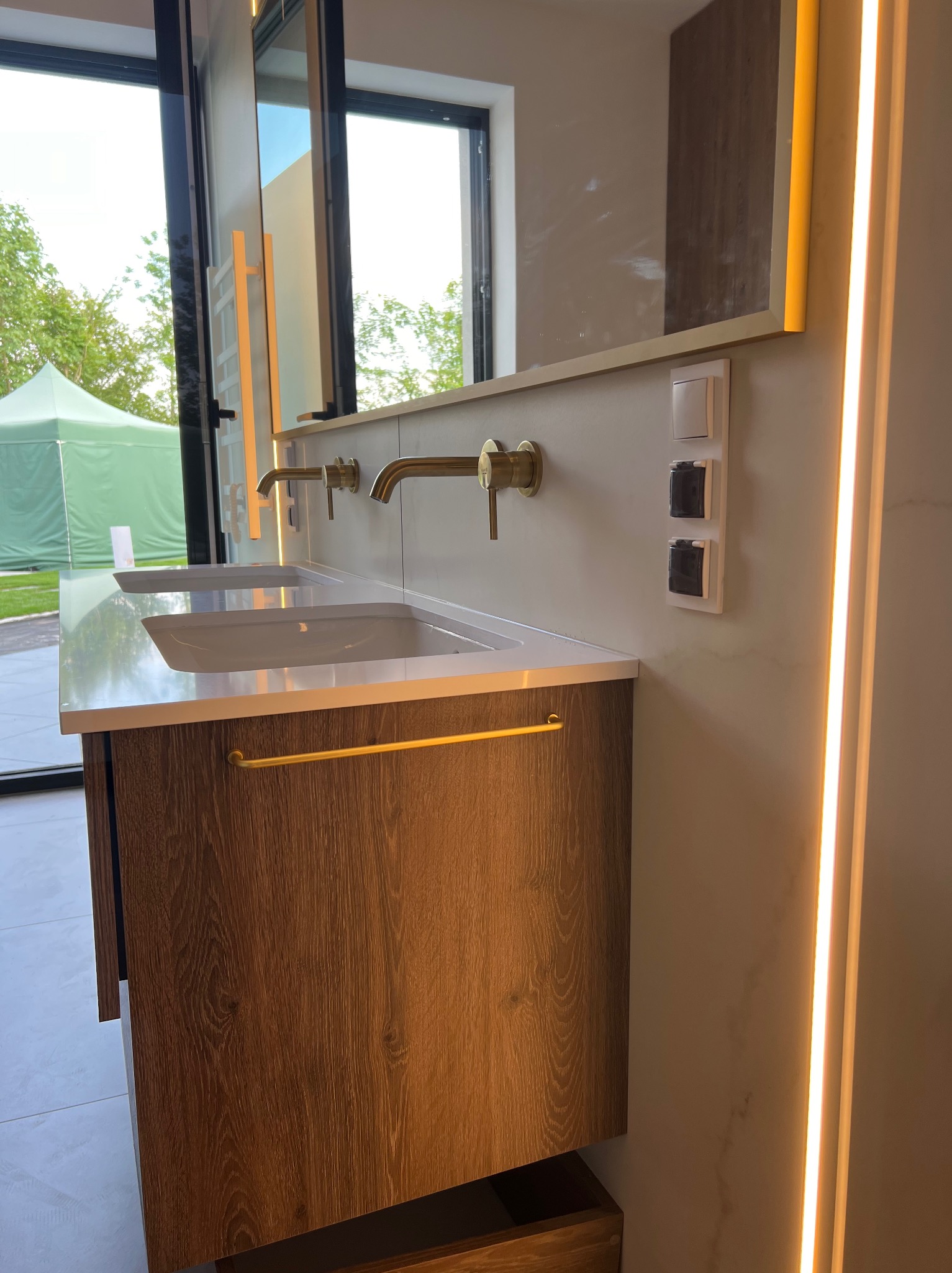 Nowoczesna łazienka z podwójną umywalką nablatową, złotą armaturą i oświetleniem LED, widok z boku na tle okna z widokiem na ogród.