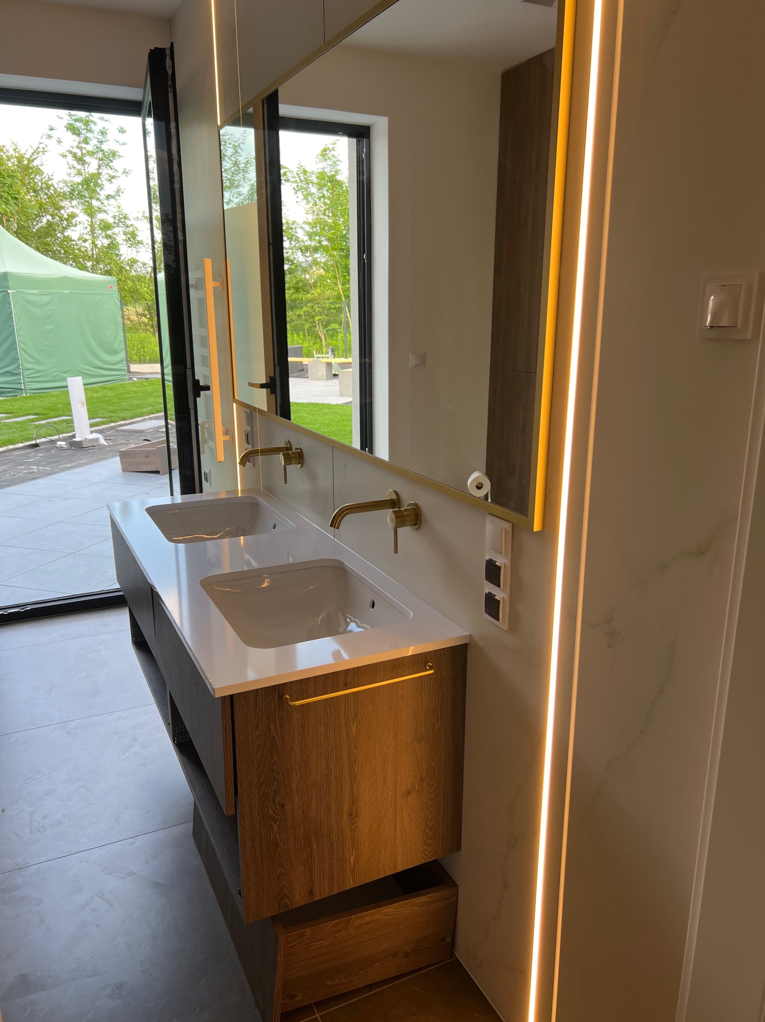 Nowoczesna łazienka z podwójną umywalką na drewnianej szafce, złotą armaturą i podświetlanym lustrem, widok na ogród przez duże okno.