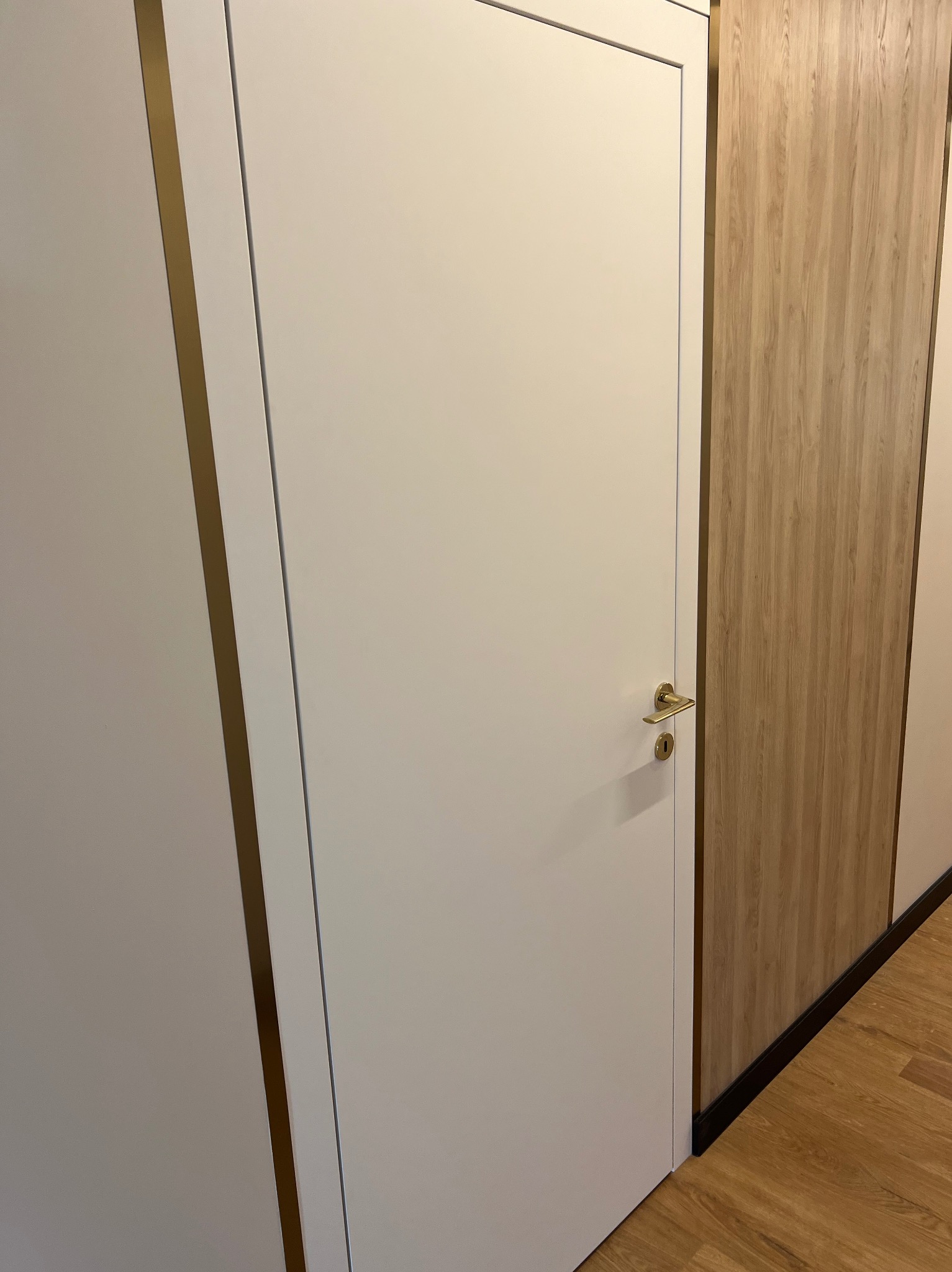 Białe drzwi wewnętrzne z delikatną złotą klamką i ramą, sąsiadujące z panelem ściennym o wzorze jasnego drewna, uzupełnione brązową listwą przypodłogową.