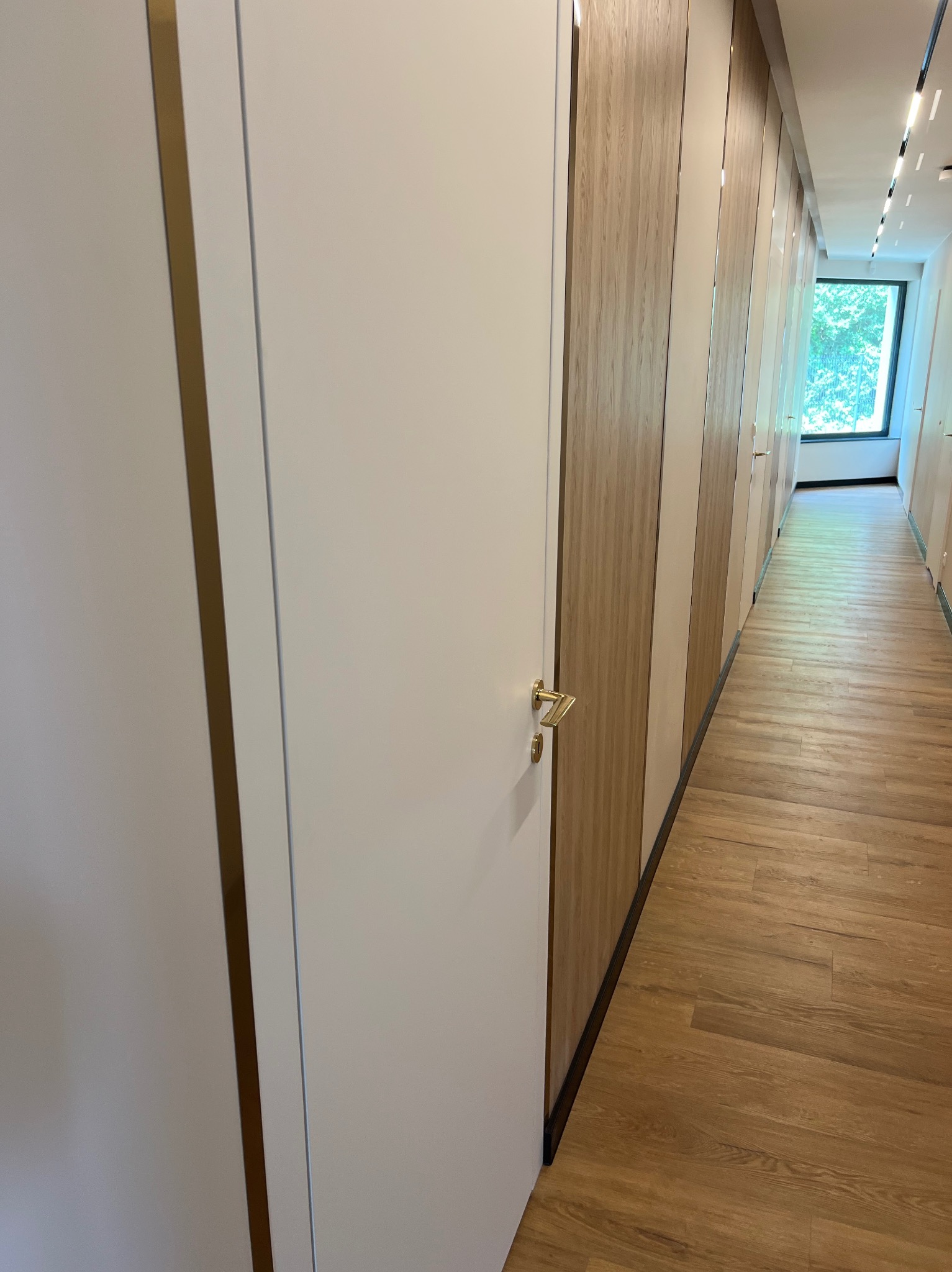 Długi korytarz z drewnianą podłogą i szeregiem białych drzwi z złotymi klamkami, przechodzący w jasne, przeszklone zakończenie z widokiem na zieleń.