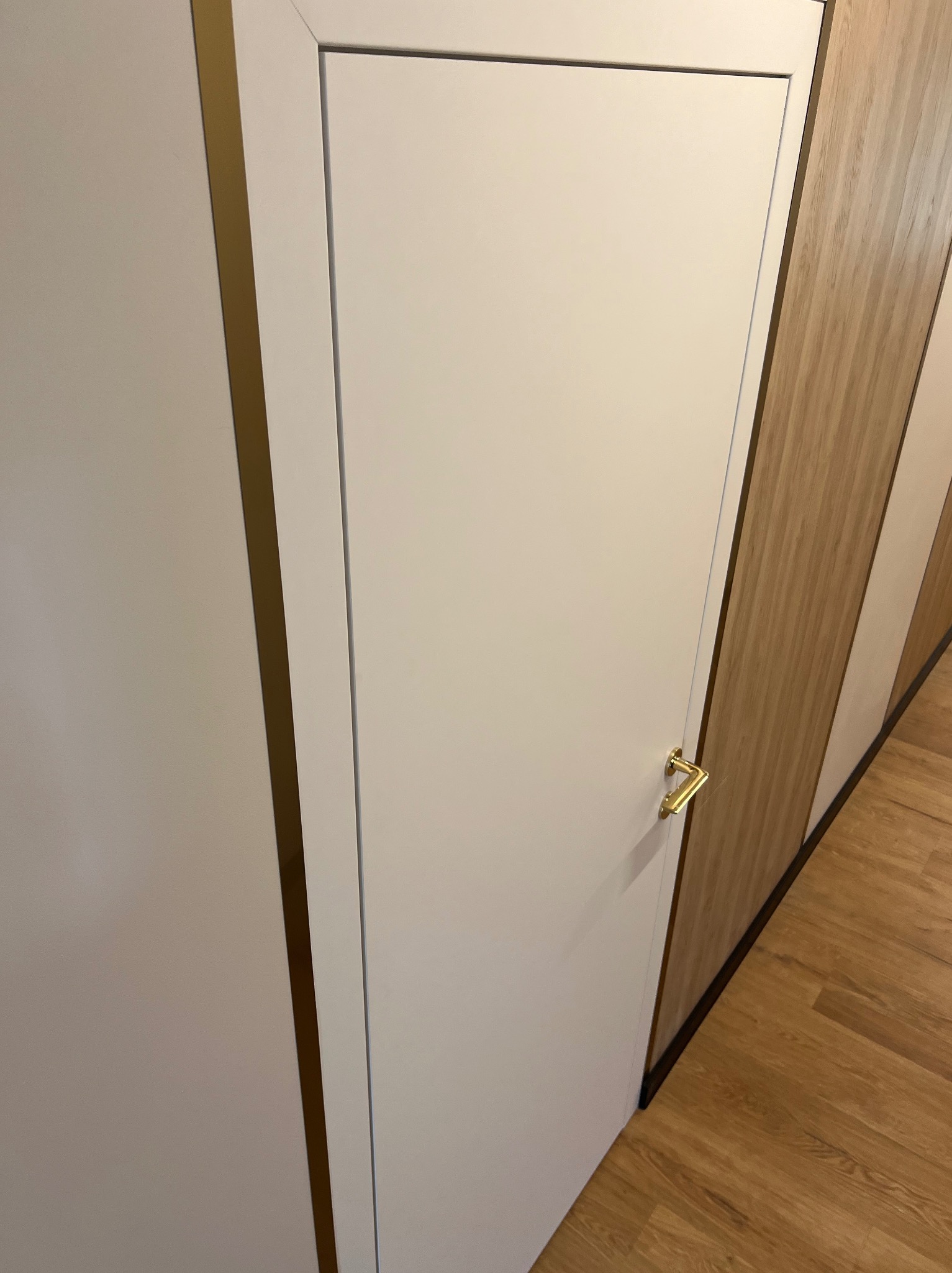 Białe, minimalistyczne drzwi wewnętrzne z złotą klamką, widoczne na tle drewnianej podłogi i zabudowy meblowej z elementami w kolorze drewna.