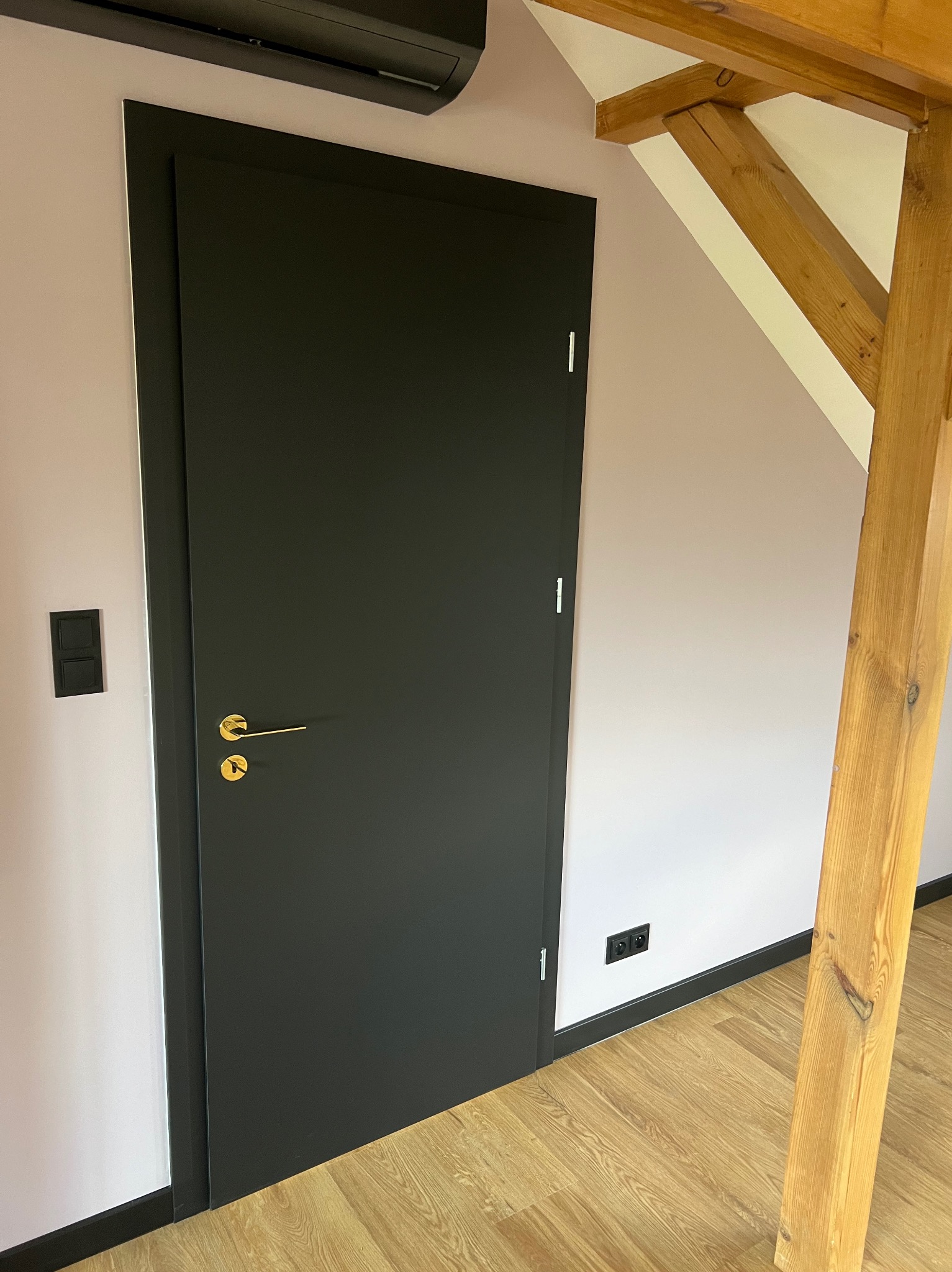 Czarna, matowa, bezprzylgowa drzwi z złotą klamką na tle jasnoróżowej ściany z drewnianą belką stropową i czarnymi listwami przypodłogowymi oraz gniazdkami, widoczny klimatyzator.