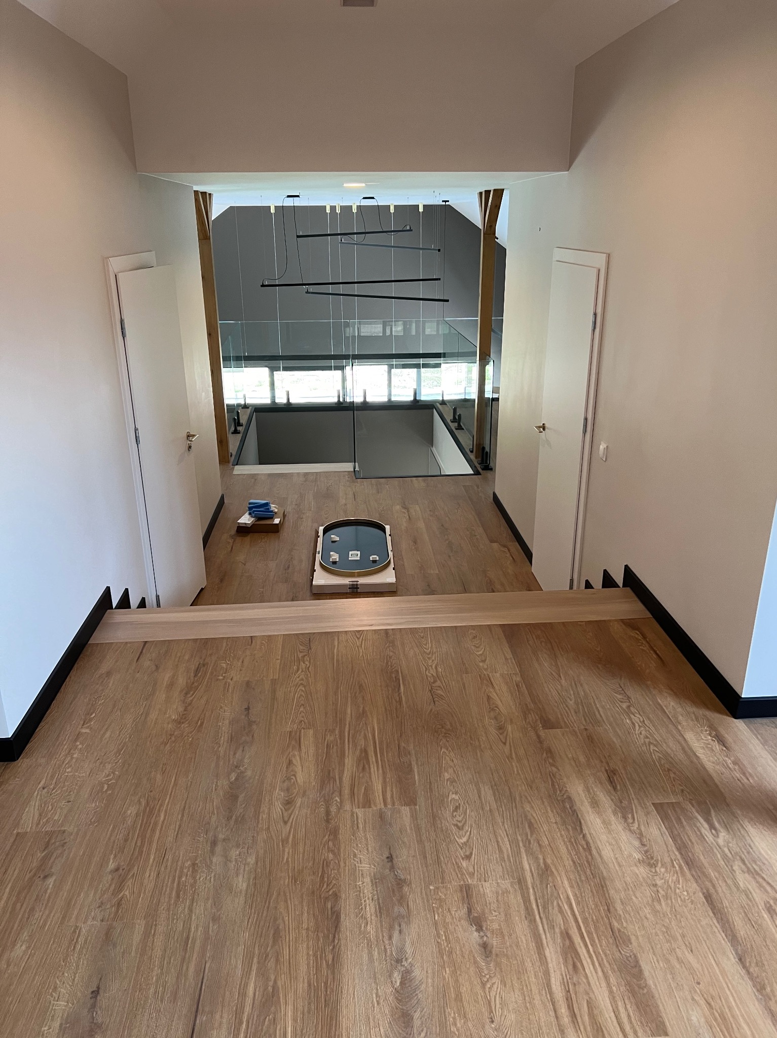 Widok z góry na korytarz z drewnianą podłogą i schodami, białe ściany z czarnymi listwami przypodłogowymi, drzwi po obu stronach, lustro w ozdobnej ramie leżące na podłodze, nowoczesne oświetlenie...