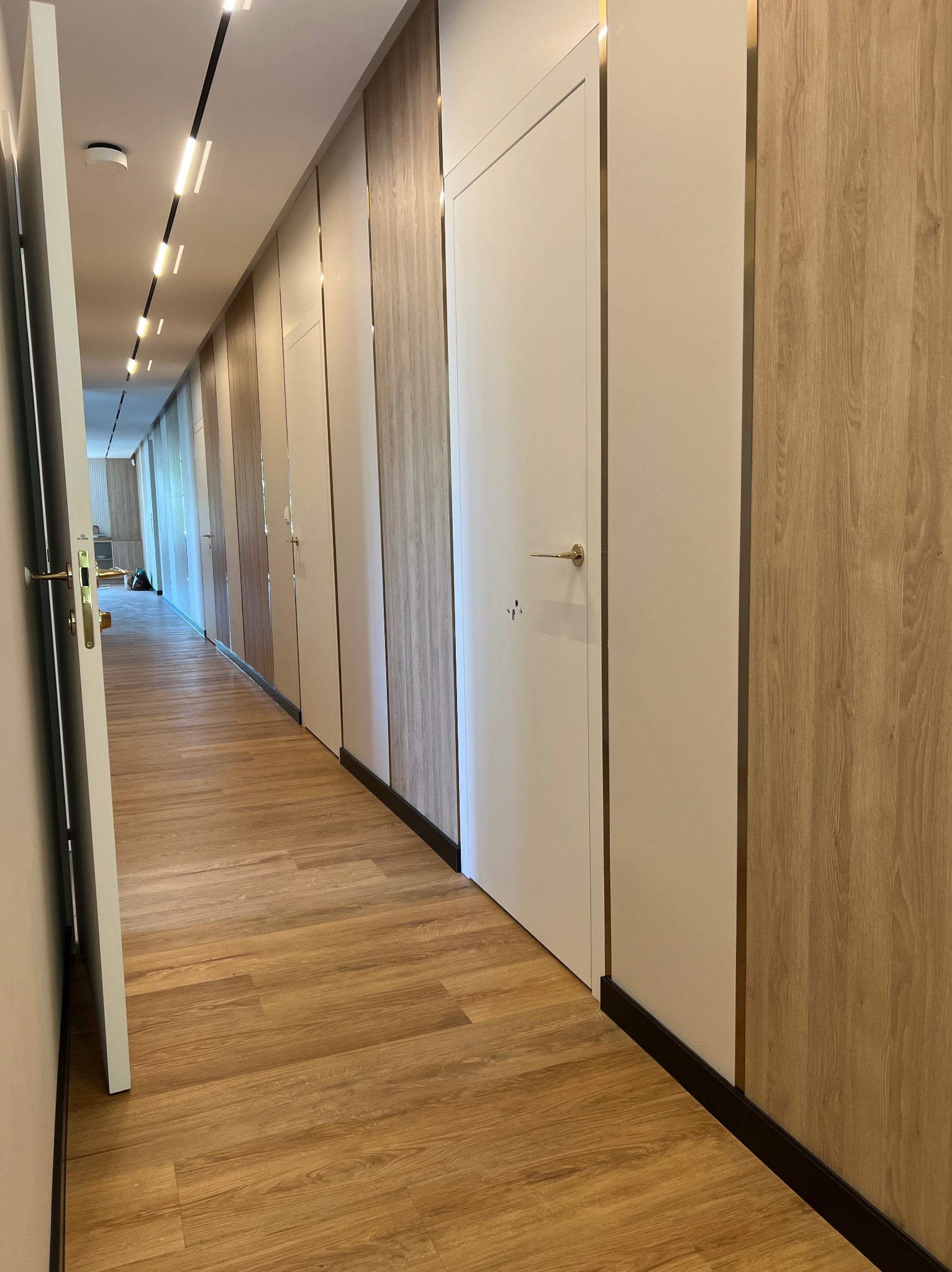 Długi, jasny korytarz z drewnopodobną podłogą i naprzemiennie białymi i drewnianymi panelami ściennymi z metalowymi akcentami, oświetlony liniowymi lampami sufitowymi, z otwartymi drzwiami na końcu...