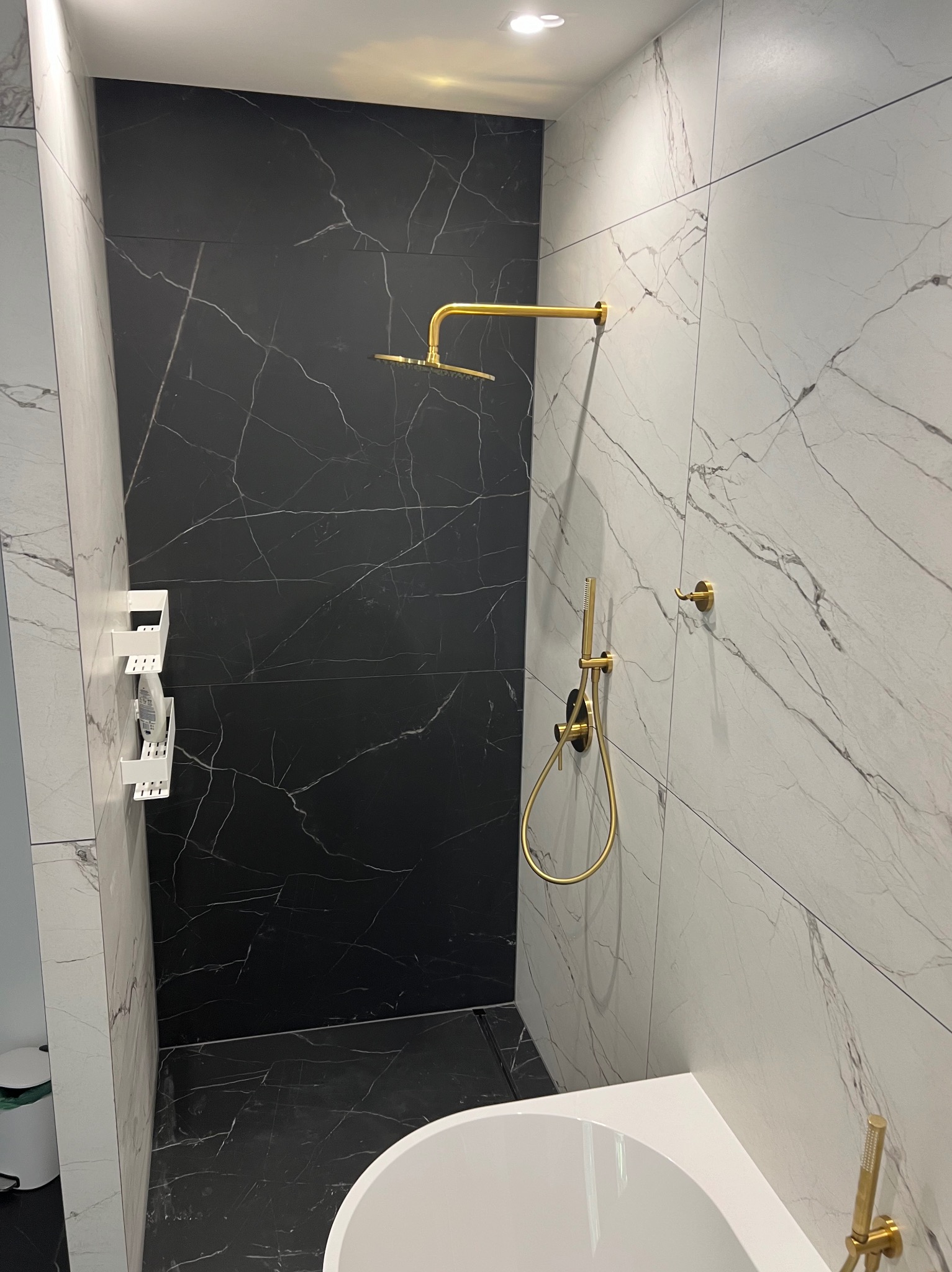 Nowoczesna łazienka z czarno-białymi marmurkowymi płytkami, złotą armaturą prysznicową i minimalistycznymi półkami na kosmetyki.