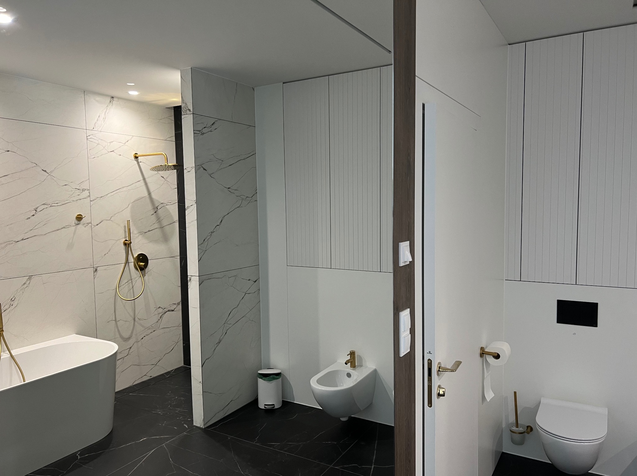 Nowoczesna łazienka z marmurowymi płytkami, złotą armaturą, wolnostojącą wanną, bidetem i podwieszaną toaletą, utrzymana w bieli i czerni.
