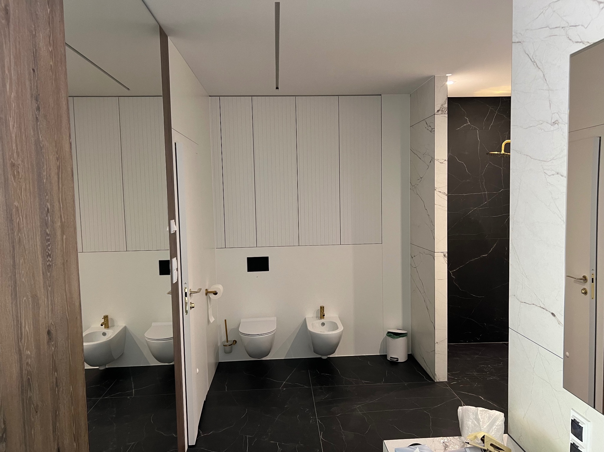 Nowoczesna łazienka z czarno-białymi marmurowymi płytkami, białymi sanitariatami z złotymi akcentami i minimalistycznymi białymi szafkami.