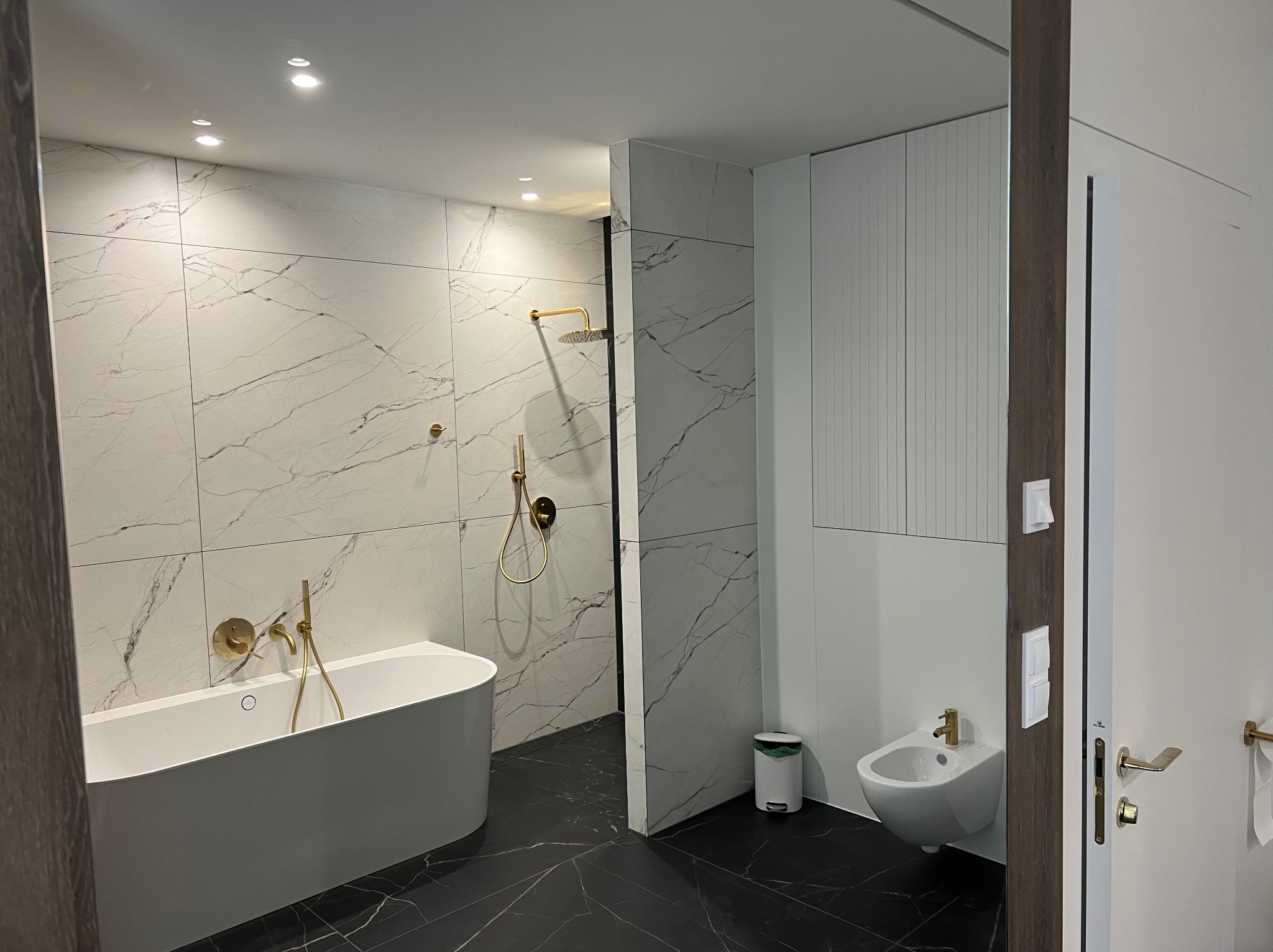 Nowoczesna łazienka z białą wanną wolnostojącą, złotą armaturą i marmurowymi płytkami na ścianach, uzupełniona o bidet i minimalistyczne szafki.