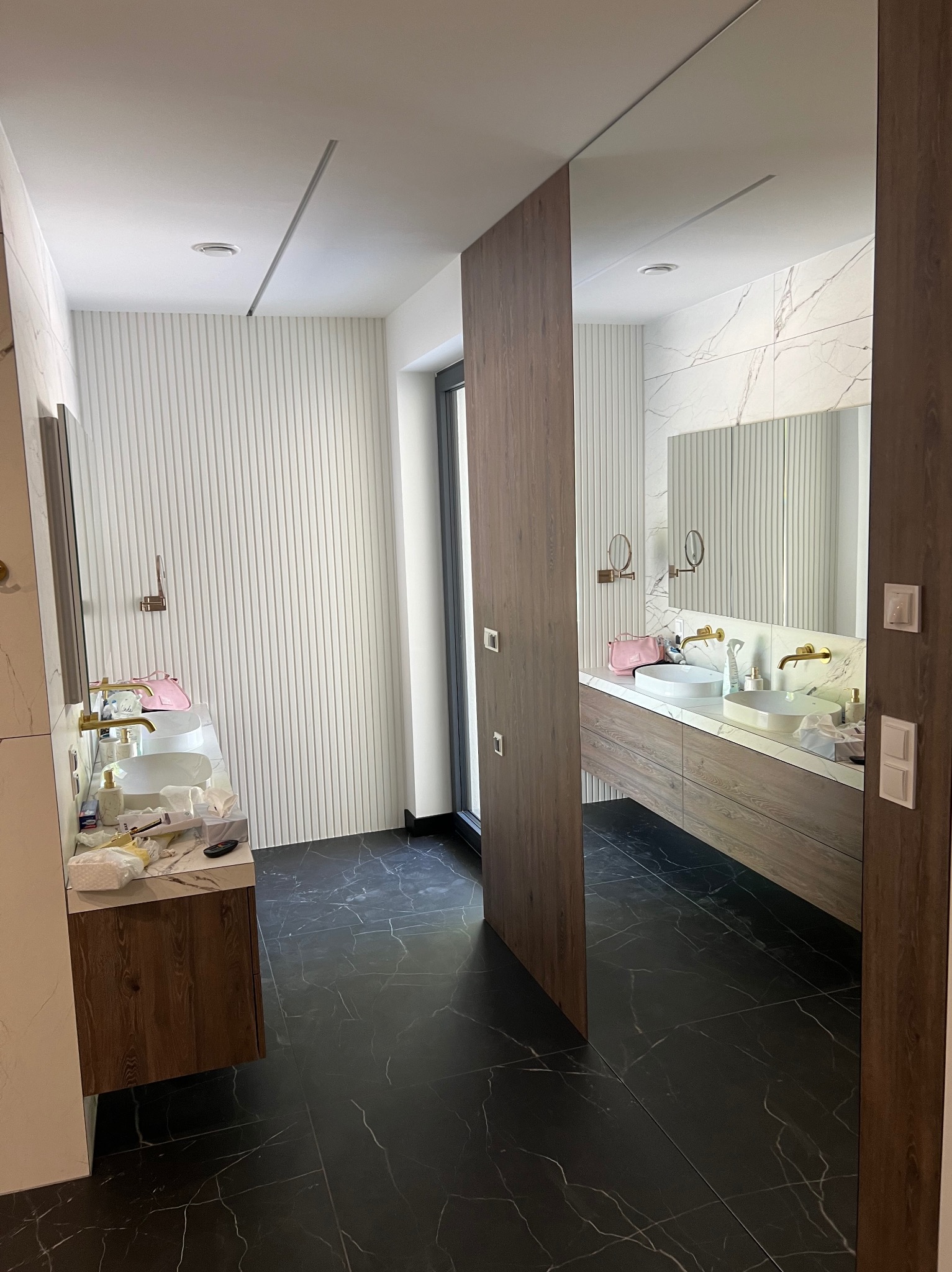 Nowoczesna łazienka z czarną marmurową podłogą, drewnianymi szafkami pod umywalkami i białymi ścianami z panelami pionowymi, widok przez otwarte drzwi.