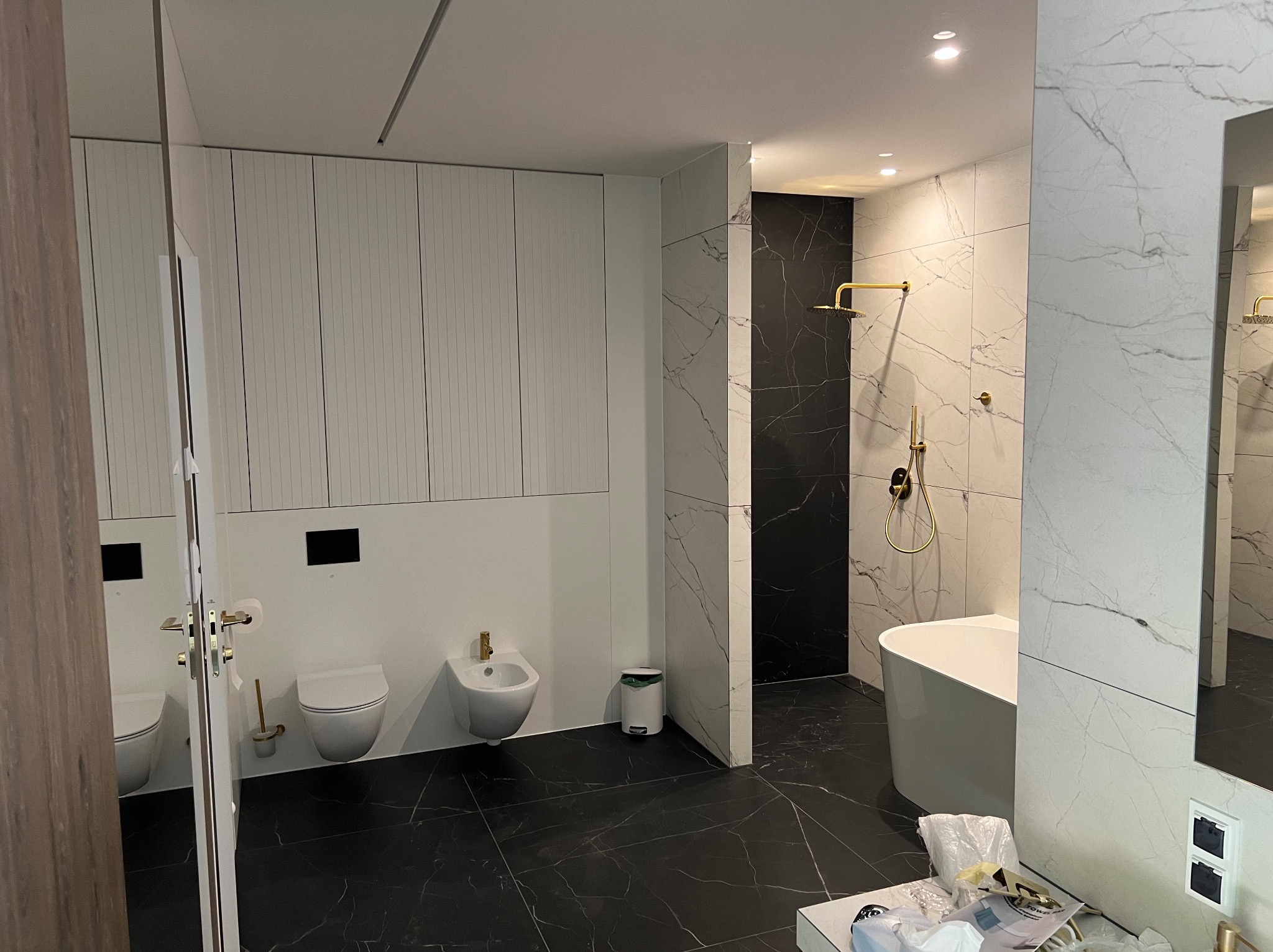 Nowoczesna łazienka z czarno-białymi marmurowymi płytkami, złota armatura, biała wolnostojąca wanna i toaleta z bidetem, minimalistyczne szafki w tle.