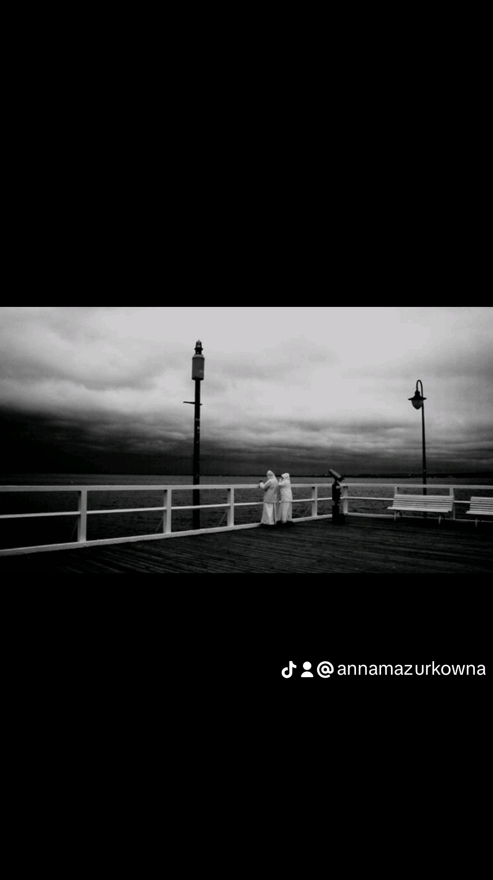 Dwie osoby w białych kombinezonach stoją na drewnianym molo, patrząc na morze pod ciemnym, burzowym niebem. Widoczna latarnia i ławki w tle.