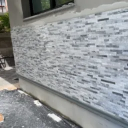 Fragment elewacji budynku z częściowo ułożoną dekoracyjną okładziną z płytek imitujących kamień, zabezpieczony folią malarską.