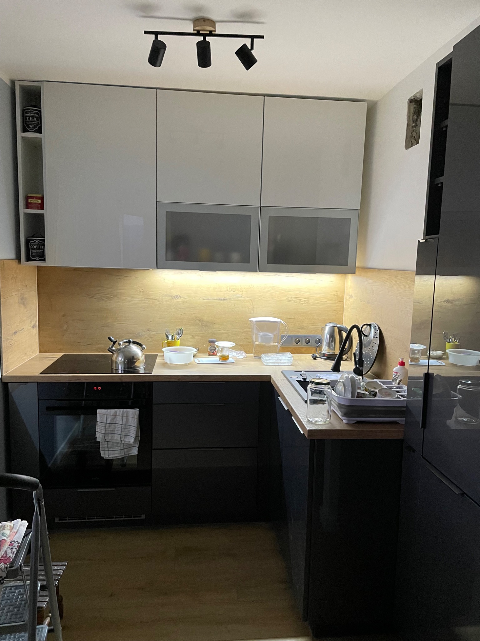 Nowoczesna kuchnia z białymi szafkami górnymi, szarymi frontami i drewnianym blatem, wyposażona w czarny piekarnik, płytę indukcyjną i zlewozmywak z suszarką. Na blacie widoczne naczynia...