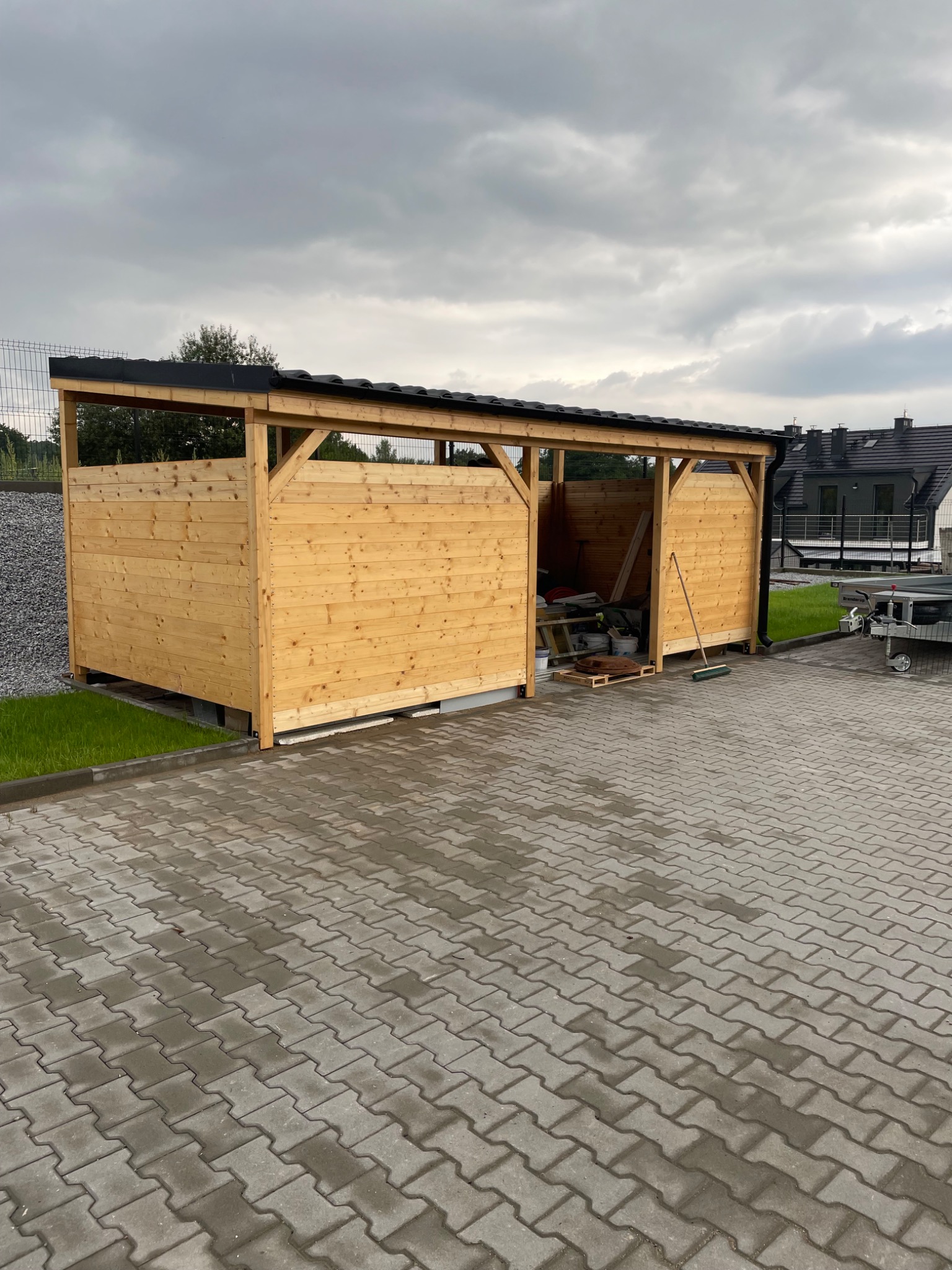 Drewniana wiata garażowa z czarnym dachem, widoczna na tle kostki brukowej i pochmurnego nieba. Wewnątrz wiata częściowo wypełniona narzędziami i akcesoriami.