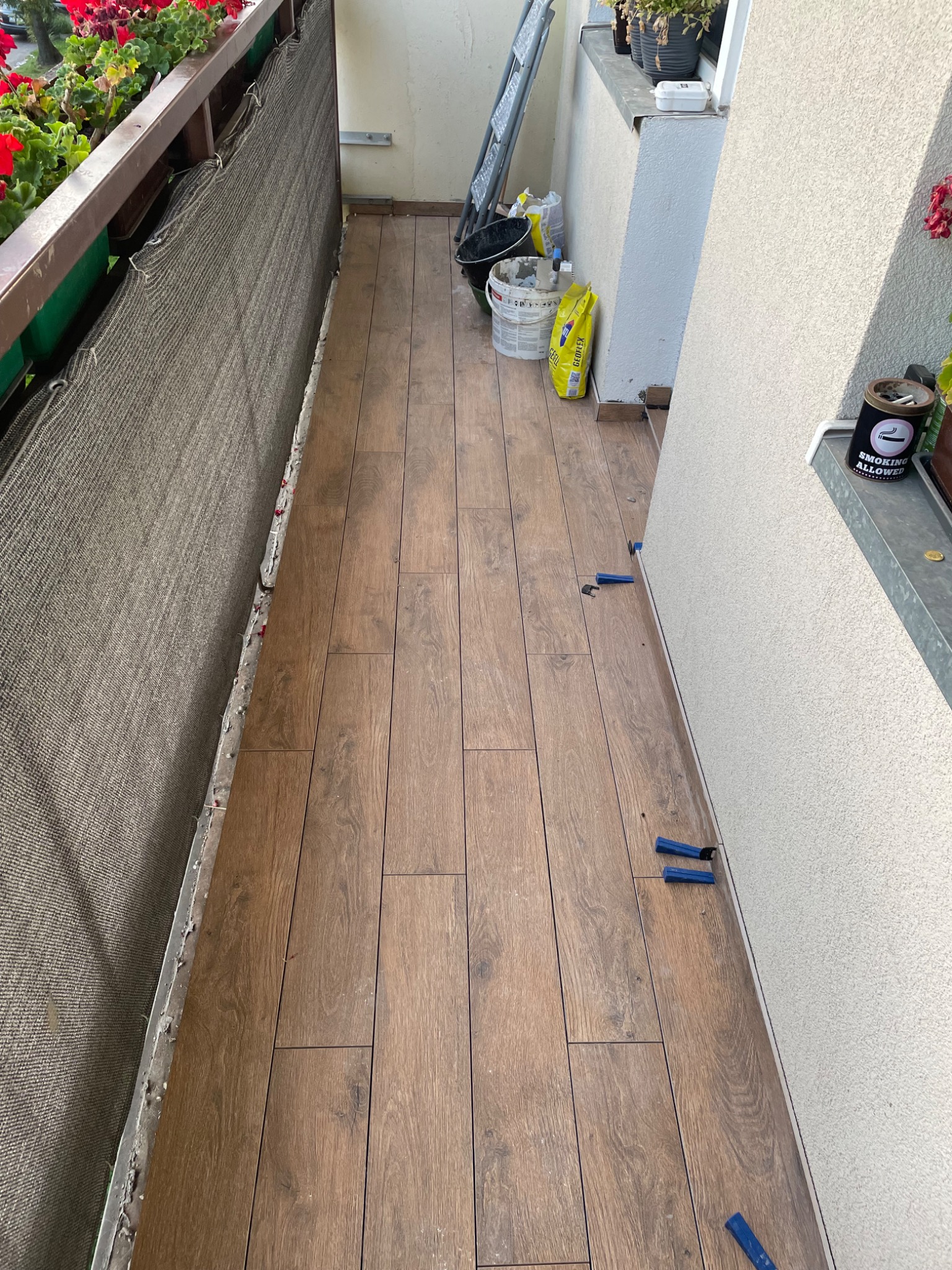 Balkon z brązowymi panelami podłogowymi ułożonymi wzdłuż, widoczne narzędzia do poziomowania płytek przy ścianie, siatka osłonowa na balustradzie i donice z czerwonymi kwiatami.