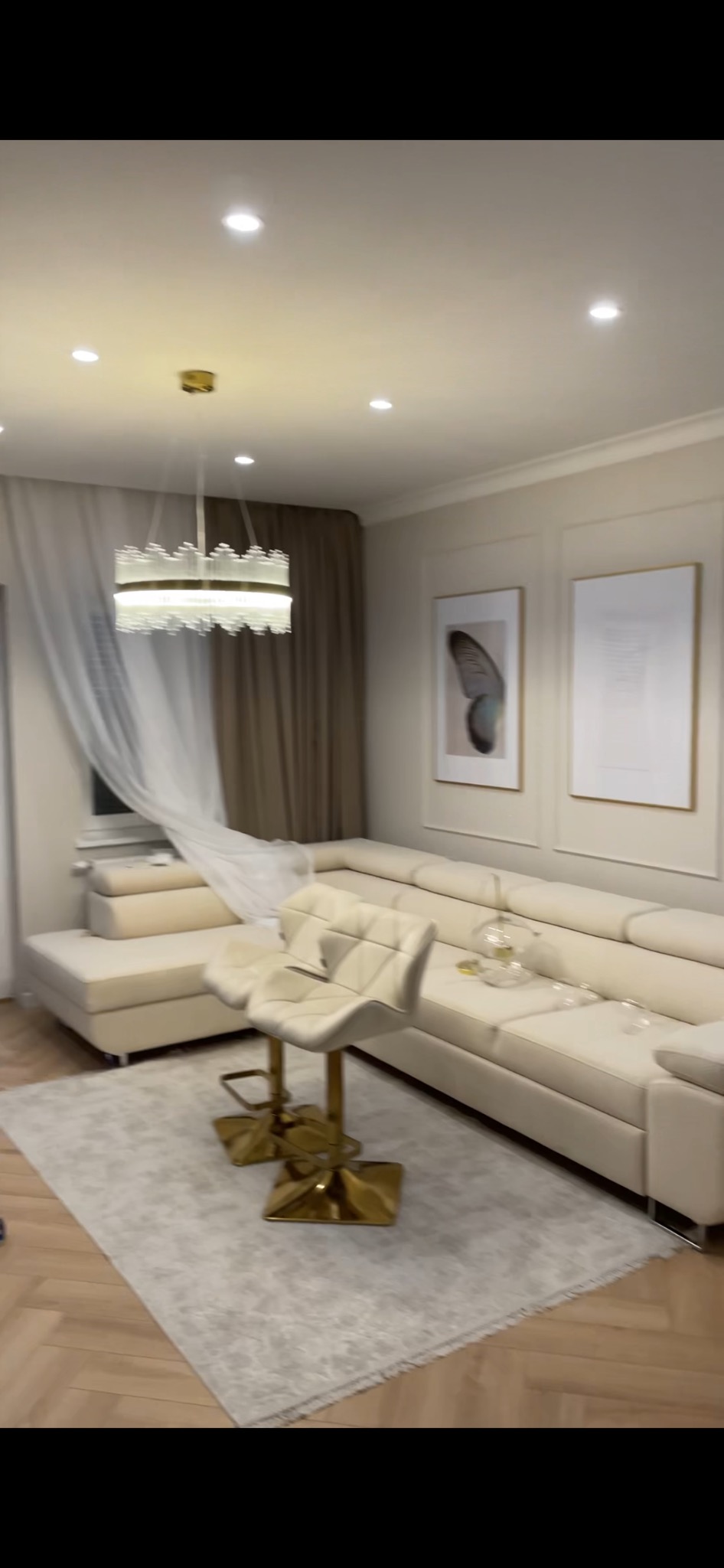 Jasny salon z narożnikiem, złotym krzesłem barowym, dywanem i dekoracyjnym oświetleniem. W tle zasłony i obrazy na ścianie.