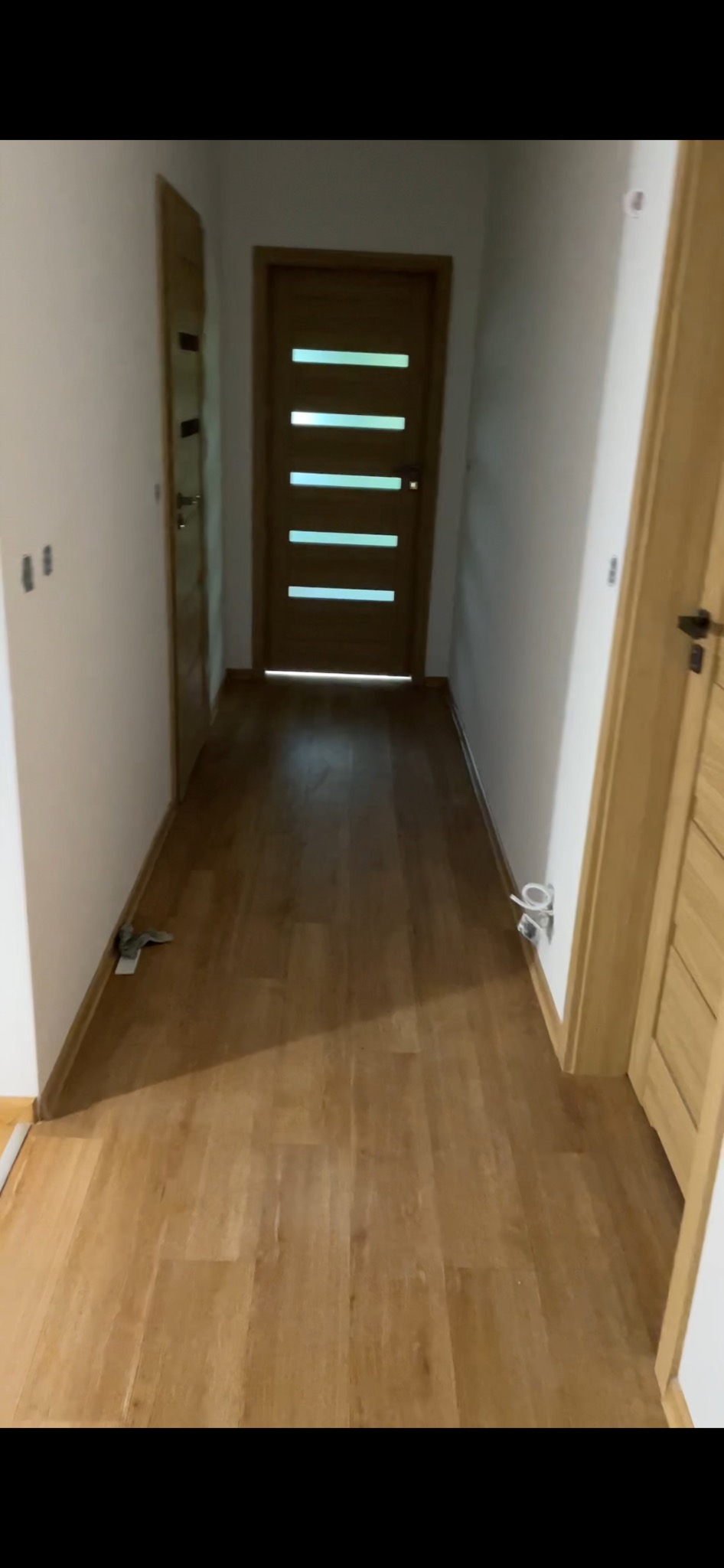 Korytarz z nową podłogą z paneli w odcieniach drewna, widoczne dwie pary drzwi po obu stronach, ściany pomalowane na biało, na podłodze leżą drobne śmieci budowlane.