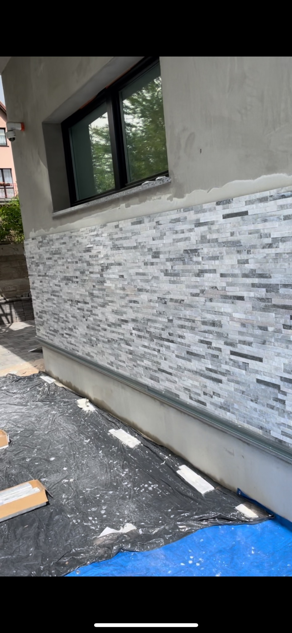 Fragment elewacji budynku z częściowo ułożoną dekoracyjną okładziną z płytek imitujących kamień, zabezpieczony folią malarską.