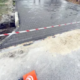Świeżo ułożona, mokra, szara kostka brukowa, oddzielona taśmą ostrzegawczą od piasku, z pomarańczowym pachołkiem na pierwszym planie i pracownikami w oddali.