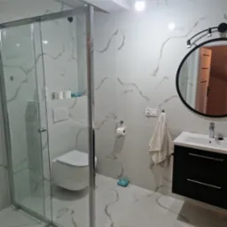 Wykończona łazienka z kabiną prysznicową ze szklanymi drzwiami, podwieszaną toaletą, czarną szafką z umywalką i okrągłym lustrem z oświetleniem punktowym, wykończona białymi płytkami imitującymi...