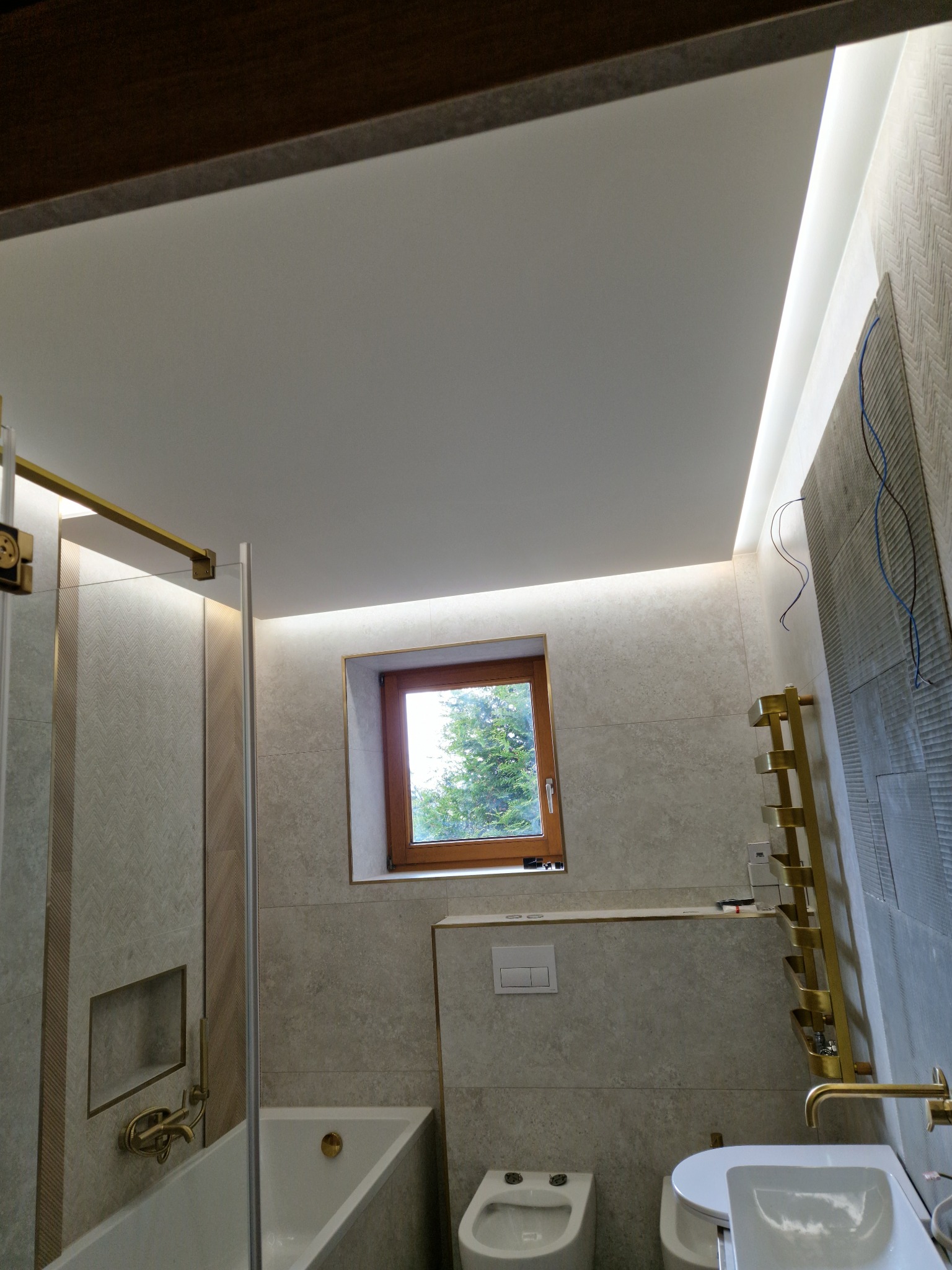 Nowoczesna łazienka w odcieniach beżu z drewnianymi akcentami, oświetleniem LED i oknem z widokiem na drzewo, złota armatura i grzejnik drabinkowy, płytki z delikatnym wzorem.