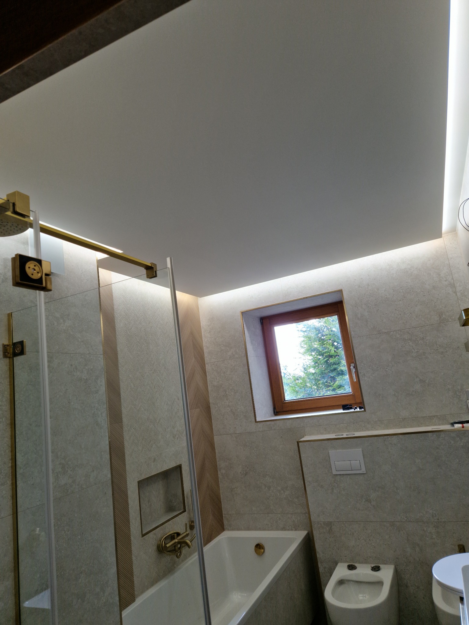 Nowoczesna łazienka z beżowymi płytkami, kabiną prysznicową ze złotymi elementami, oknem z widokiem na drzewa i podświetleniem LED.