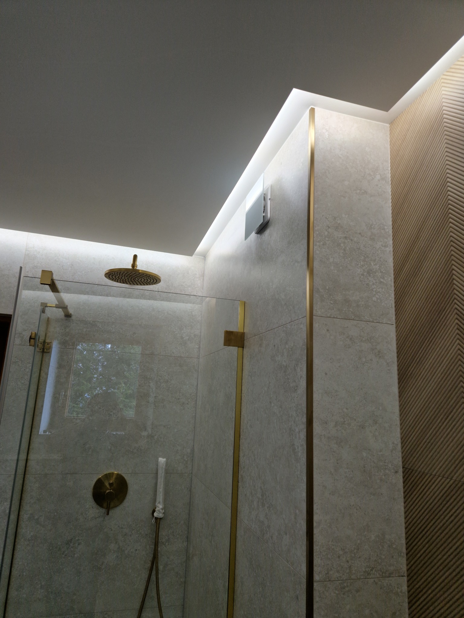 Nowoczesna łazienka z kabiną prysznicową typu walk-in, wykończona płytkami w odcieniach szarości i złotymi akcentami, w tym armatura i listwy dekoracyjne, oświetlenie LED wzdłuż sufitu.