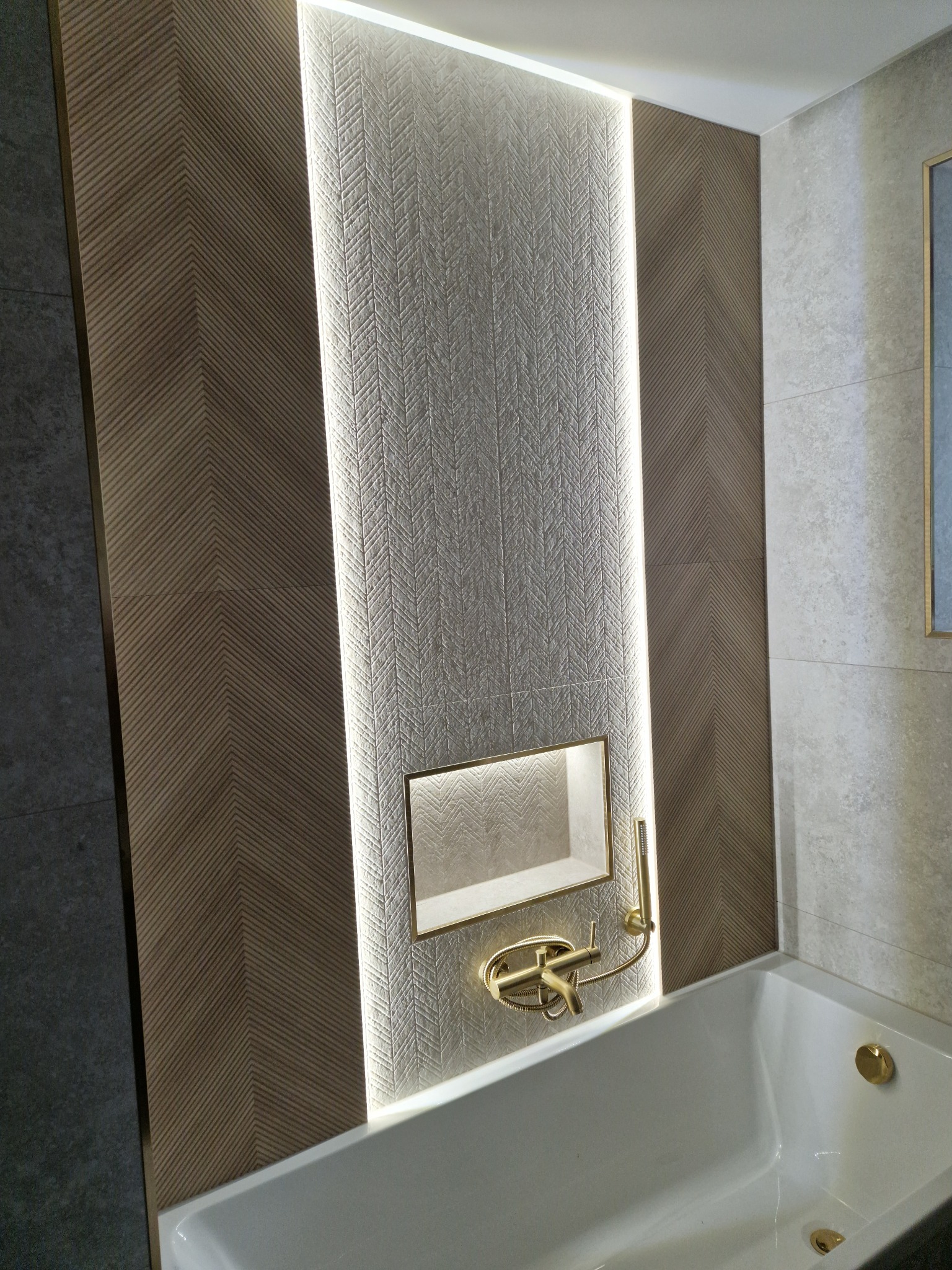 Nowoczesna łazienka z podświetloną wnęką i baterią w kolorze złotym, ściany wyłożone płytkami w geometryczny wzór jodełki.