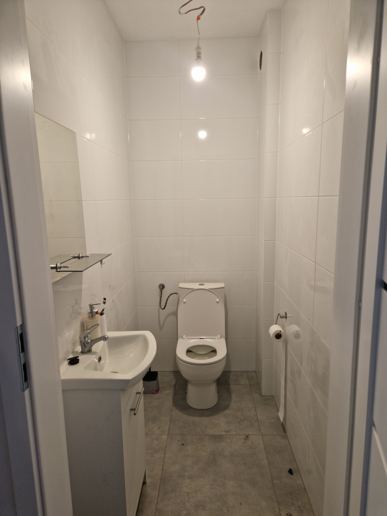 Wnętrze wykończonej łazienki z białymi płytkami na ścianach i szarą podłogą, widoczna umywalka z szafką, toaleta i wiszący papier toaletowy, z sufitu zwisa żarówka na kablu.