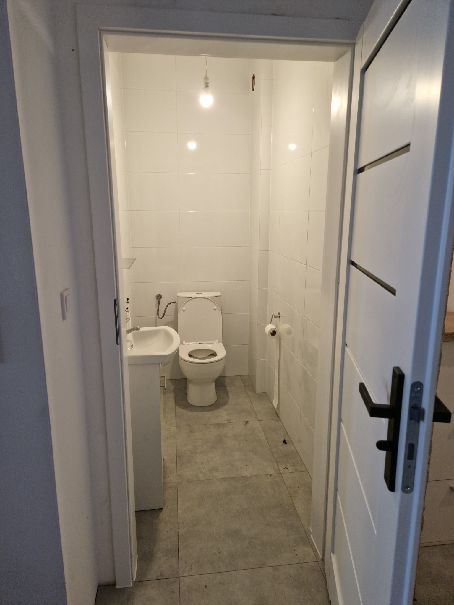 Wnętrze małej łazienki z białą ceramiką, wykończonej jasnymi płytkami, widok zza uchylonych drzwi z czarną klamką.