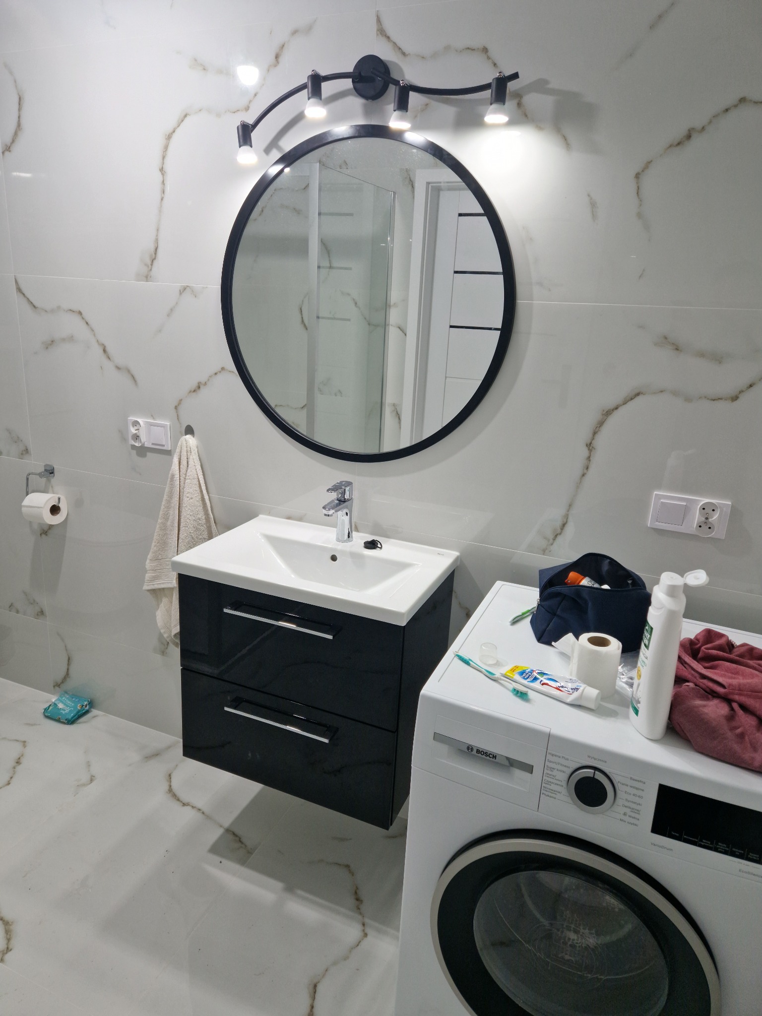 Nowoczesna łazienka z biało-złotymi płytkami imitującymi marmur, czarną szafką pod umywalką z chromowanymi uchwytami, okrągłym lustrem z czarną ramą i lampą z kilkoma punktami świetlnymi, pralką...