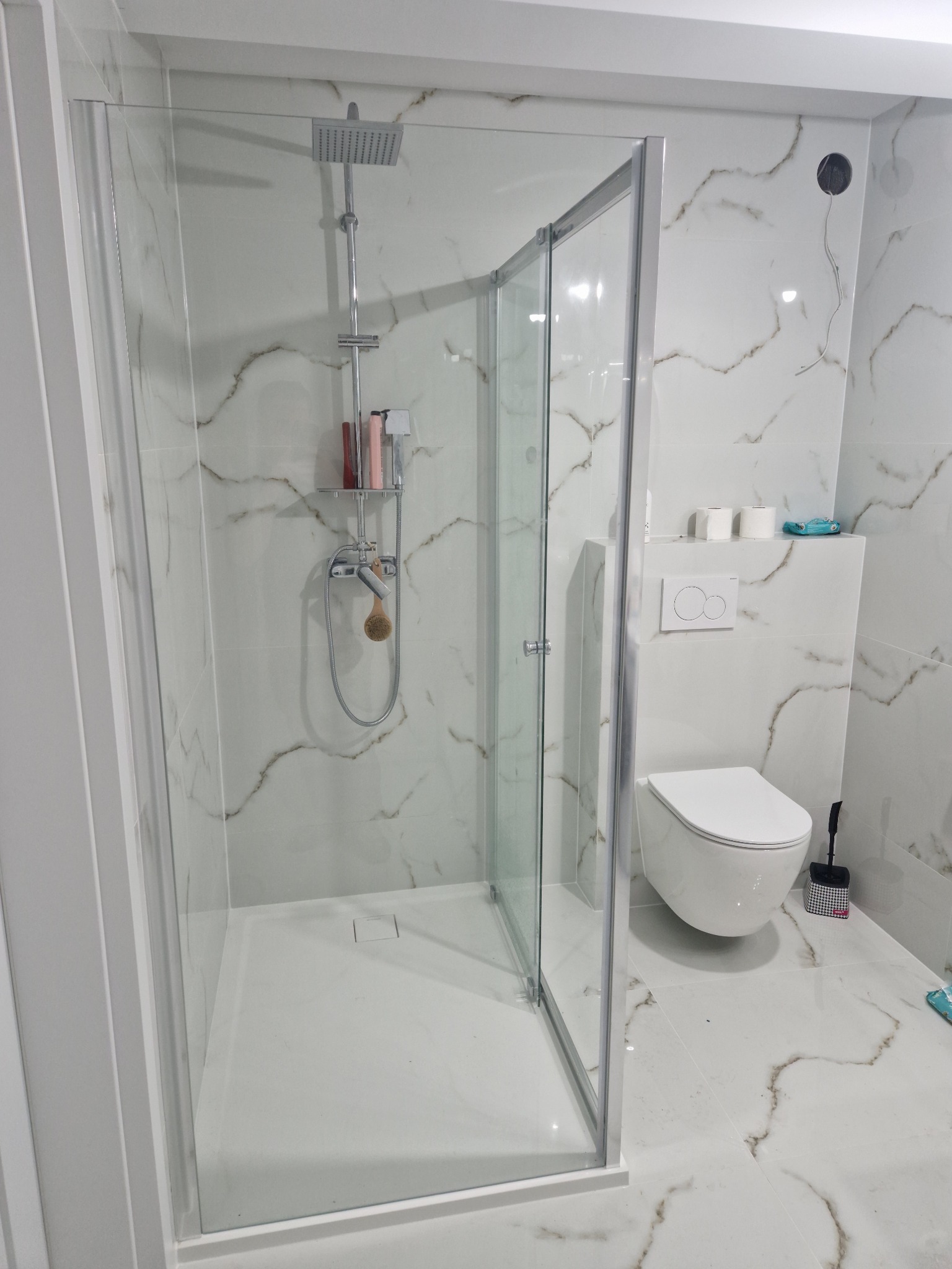 Nowoczesna kabina prysznicowa z deszczownicą, białe płytki imitujące marmur z brązowymi żyłami, wisząca toaleta, akcesoria łazienkowe w minimalistycznym wnętrzu.
