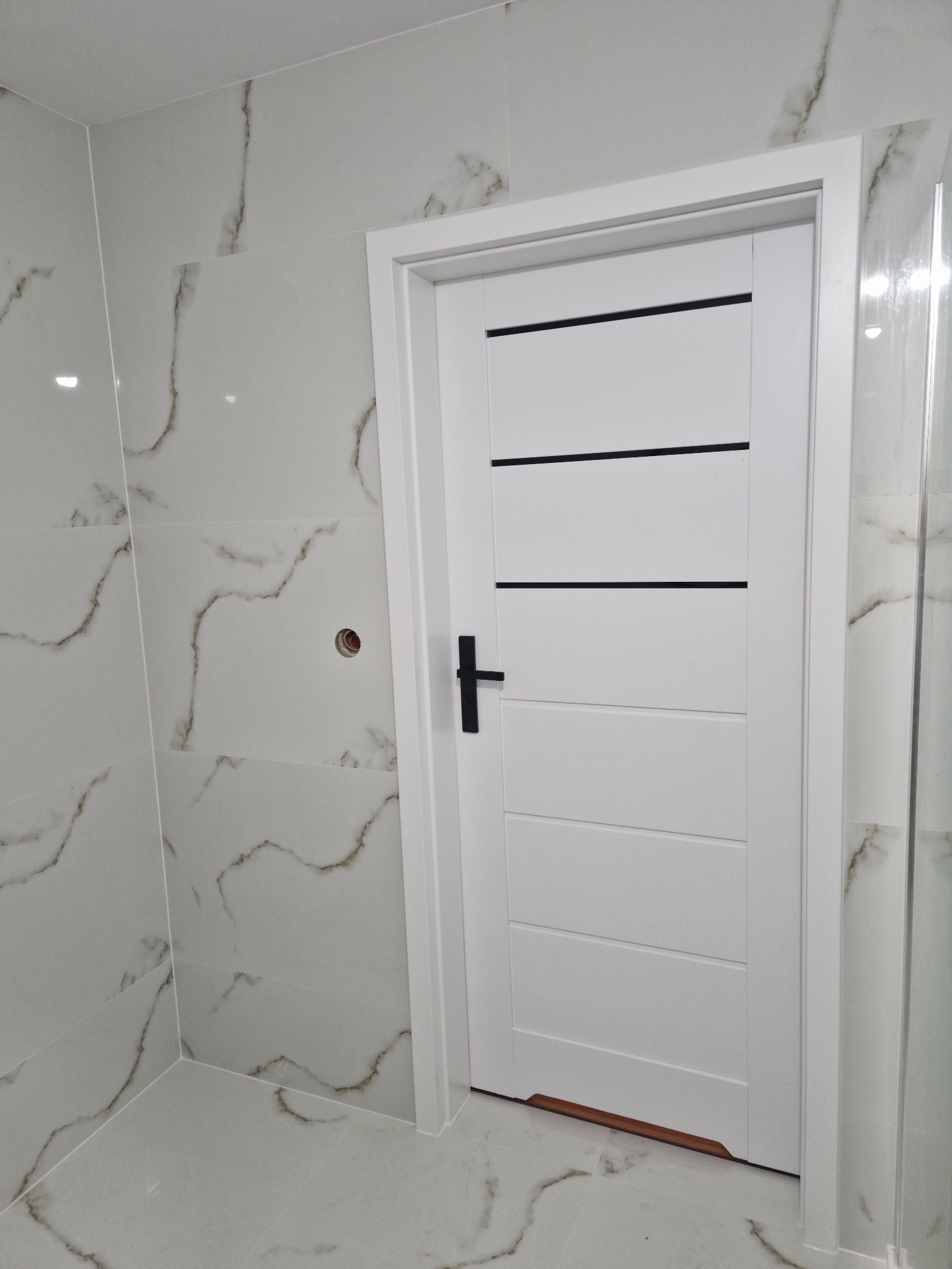 Wykończona łazienka z białymi płytkami imitującymi marmur i białą, nowoczesną stolarką drzwiową z czarnymi poziomymi akcentami, widoczny fragment kabiny prysznicowej.