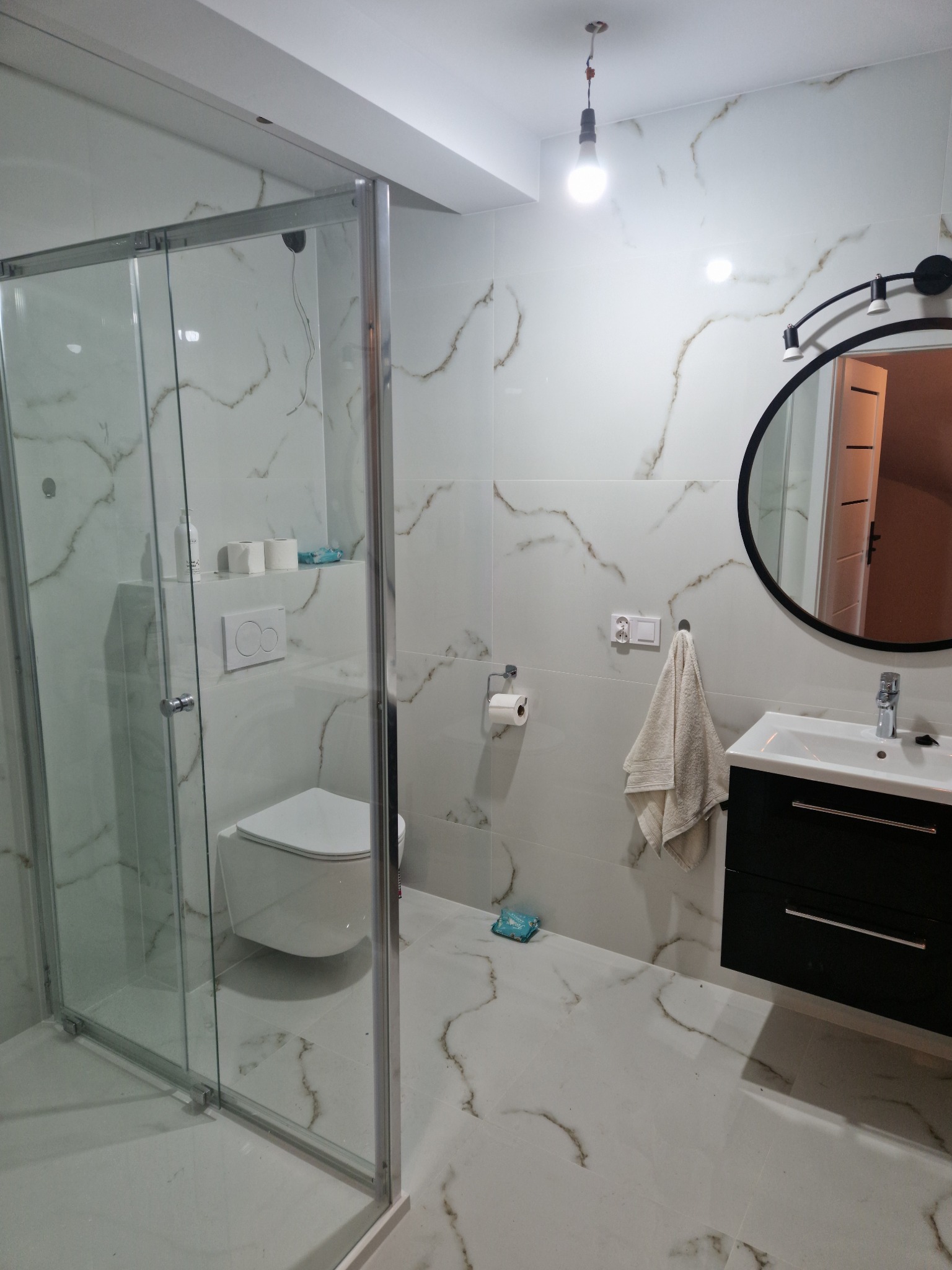 Wykończona łazienka z kabiną prysznicową ze szklanymi drzwiami, podwieszaną toaletą, czarną szafką z umywalką i okrągłym lustrem z oświetleniem punktowym, wykończona białymi płytkami imitującymi...