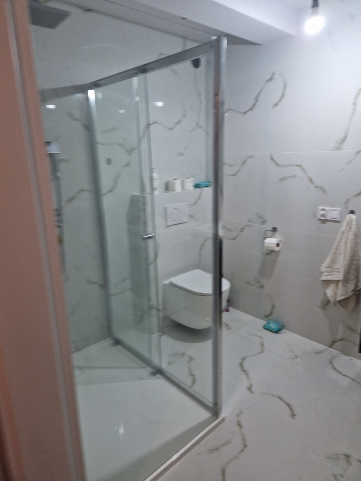 Nowoczesna łazienka z kabiną prysznicową typu walk-in, białe płytki z marmurkowym wzorem na ścianach i podłodze, wisząca toaleta, ręcznik na wieszaku, oświetlenie sufitowe.