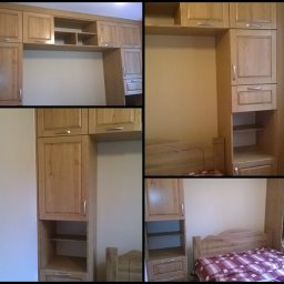 Drewnowo - Zabudowa drewniana w sypialni, obejmująca szafki wiszące nad łóżkiem i szafki boczne z półkami i szufladami, wykonana z jasnego drewna z widocznym usłojeniem i srebrnymi uchwytami.