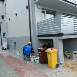 Mężczyzna w niebieskiej kurtce maluje elewację budynku na szaro, widoczne puszki z farbą, zabezpieczona kostka brukowa i dwa pojemniki na śmieci.