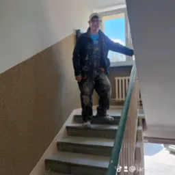 Pracownik w ubraniu roboczym, pokrytym śladami farby, stoi na schodach w klatce schodowej w Oleśnicy, gdzie ściany są częściowo pomalowane na biało, a częściowo mają fakturę.