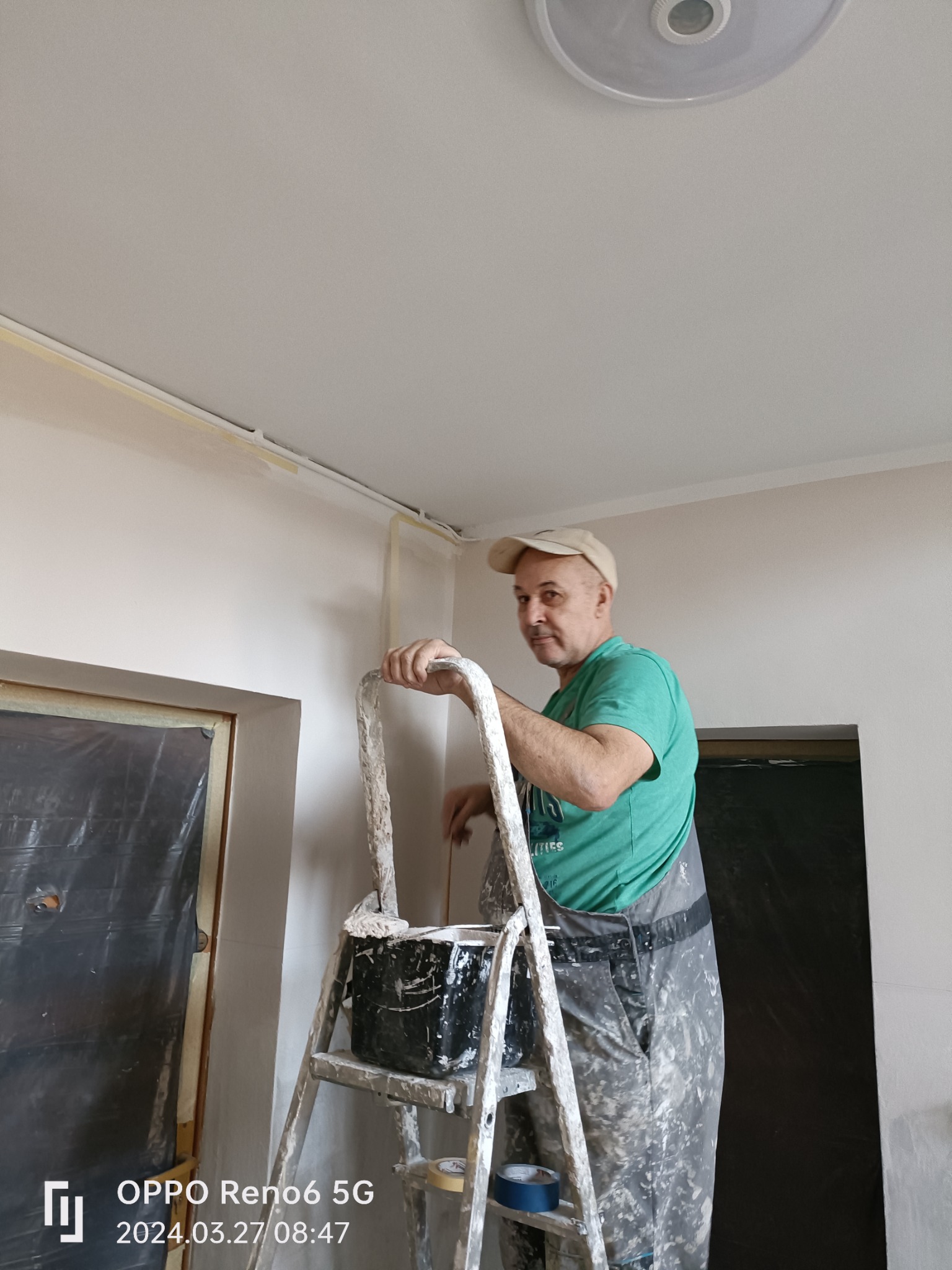Mężczyzna w ubrudzonym farbą kombinezonie i czapce stoi na drabinie z wiadrem farby, przygotowując ścianę do malowania, zabezpieczoną taśmą malarską, z widocznym fragmentem lampy sufitowej.